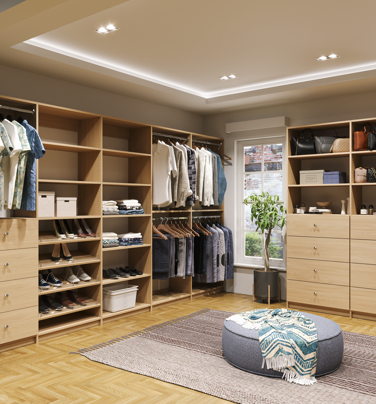 A neatly organized walk-in closet with wooden shelving units, clothing, shoes, and accessories. There's a window with a potted plant underneath and a textured area rug with a decorative pouf and a throw blanket.