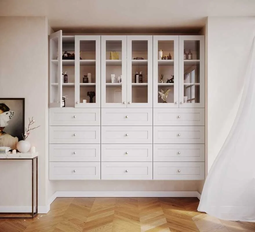 White built-in cabinet with multiple drawers and glass-front doors, displaying decorative items and bottles, in a living room with wooden parquet flooring.