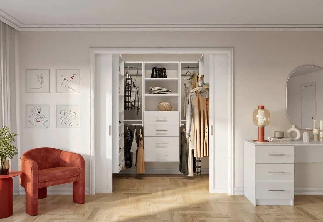 A walk-in closet with organized clothing and accessories, white shelves and drawers, and hanging clothes, framed by white molding, next to a white dresser with decorative items and a large mirror, and a small sitting area with a red velvet chair and 