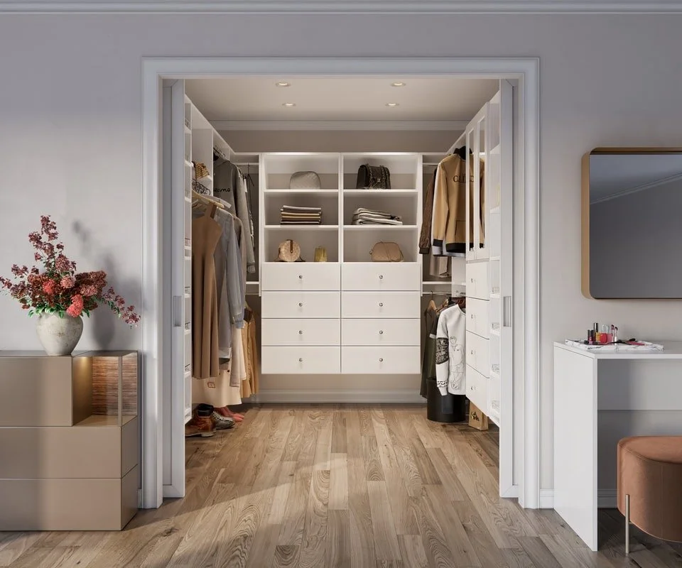 A walk-in closet with white shelving and drawers, containing neatly organized clothing, bags, and folded items. The closet is illuminated with recessed lighting. To the left, there is a beige dresser with a vase of pink flowers. On the right, a wall 