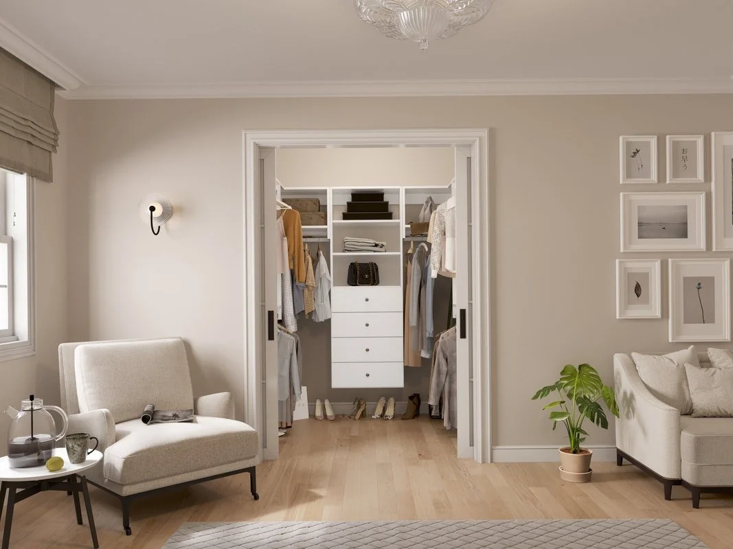 A walk-in closet with hanging clothes, shoes, and shelves, adjacent to a bedroom with seating, a coffee table, and framed artwork on the wall.