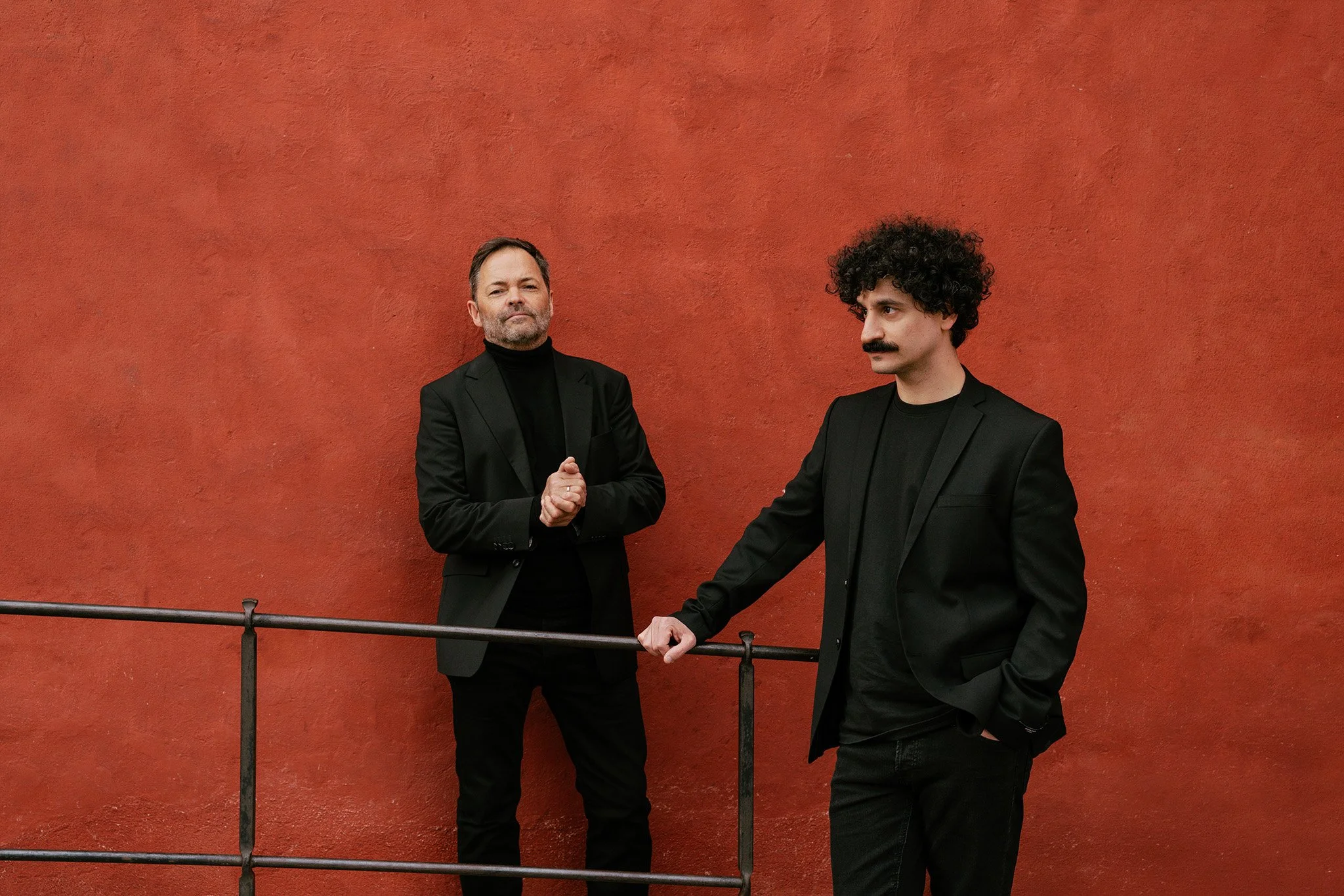 Two men dressed in black suits standing against a red wall, with one making a fist and the other holding hands.