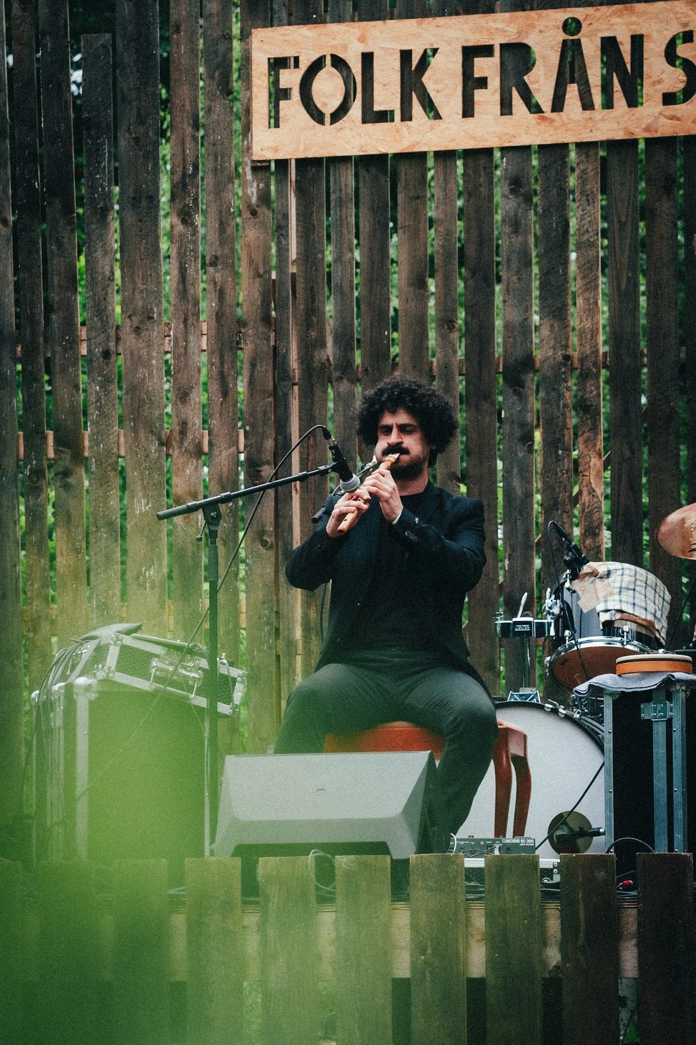 A man with curly hair and a beard playing a flute while sitting in front of a wooden fence, with musical instruments around him and a sign that reads 'FOLKERÄNS' in the background.