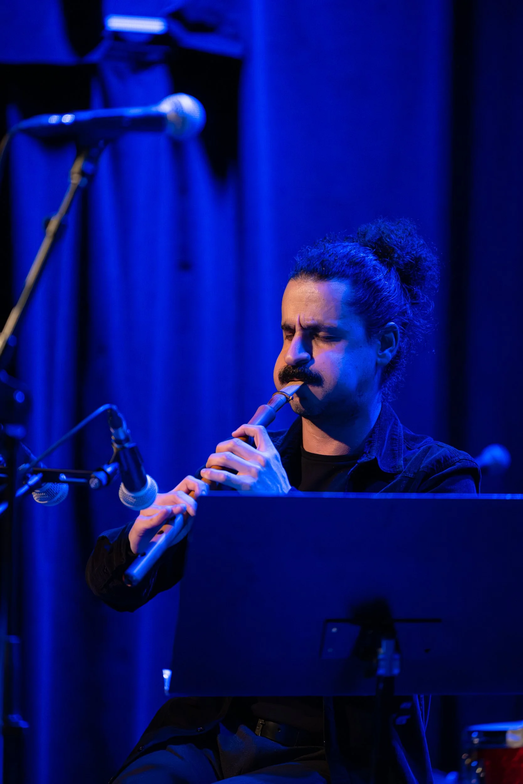 A man with dark curly hair and a mustache, wearing a black shirt, playing a wooden flute on stage under blue lighting.