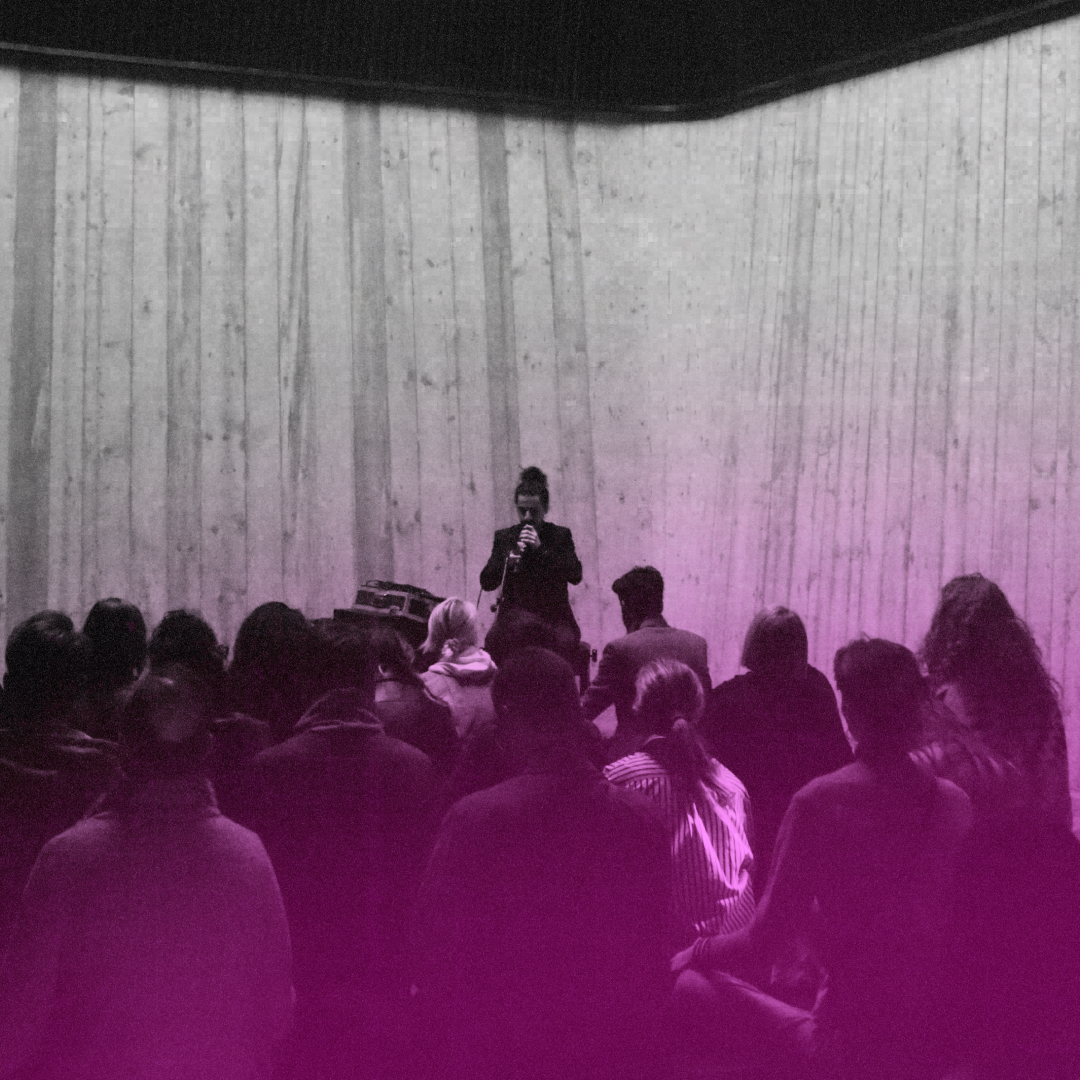 A woman speaker stands at a microphone on a stage in front of an audience in a dimly lit room with wooden wall panels, lit with purple lighting.