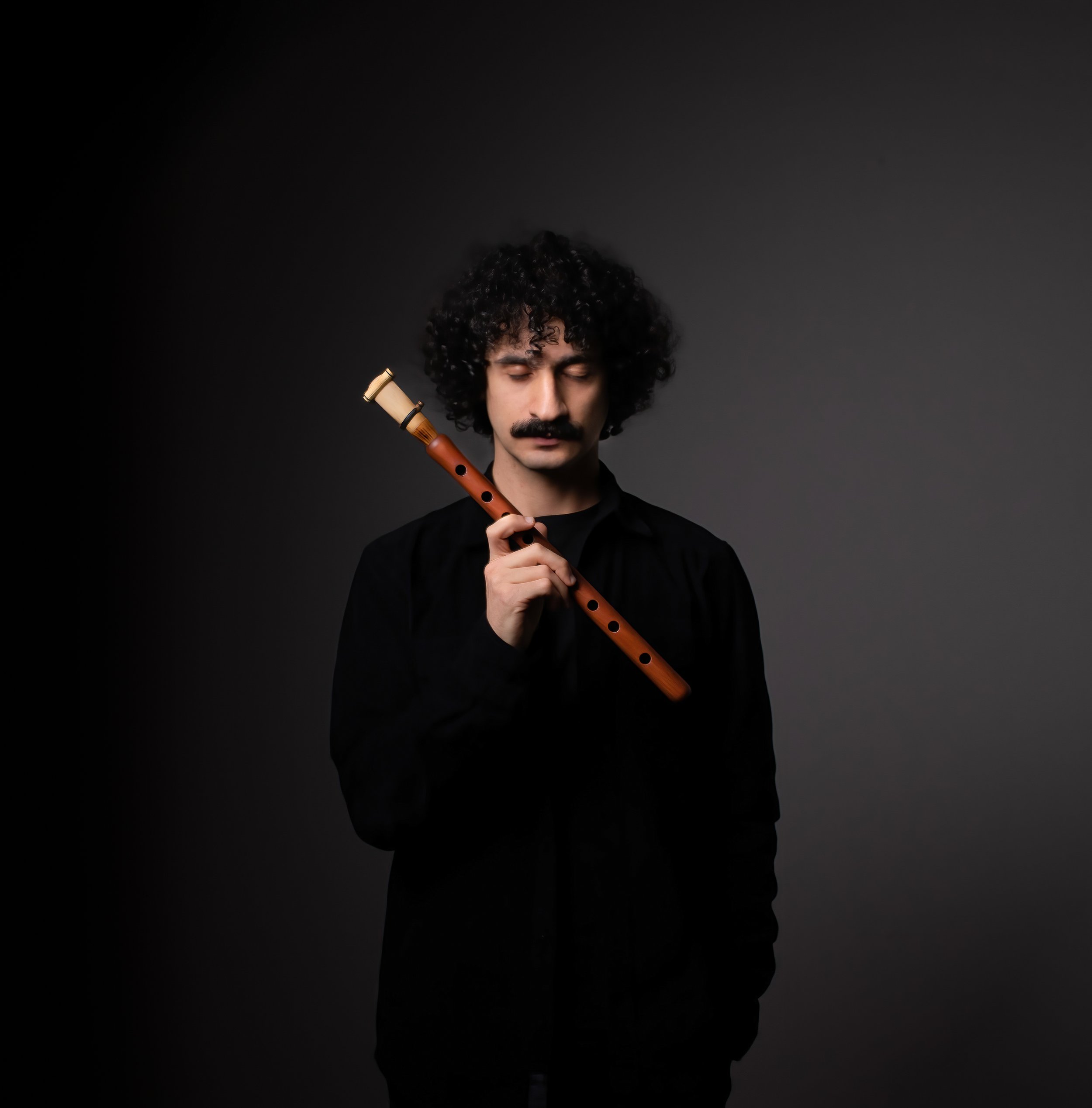 A man with curly black hair and a mustache, dressed in black, has his eyes closed while holding a wooden recorder.