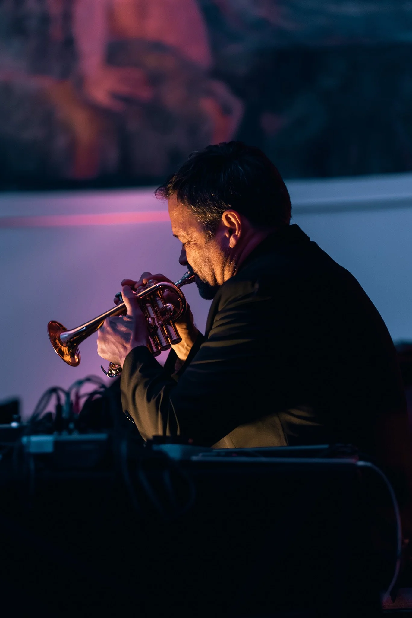 Man playing trumpet in a dimly lit room with a colorful abstract painting on the wall behind him.