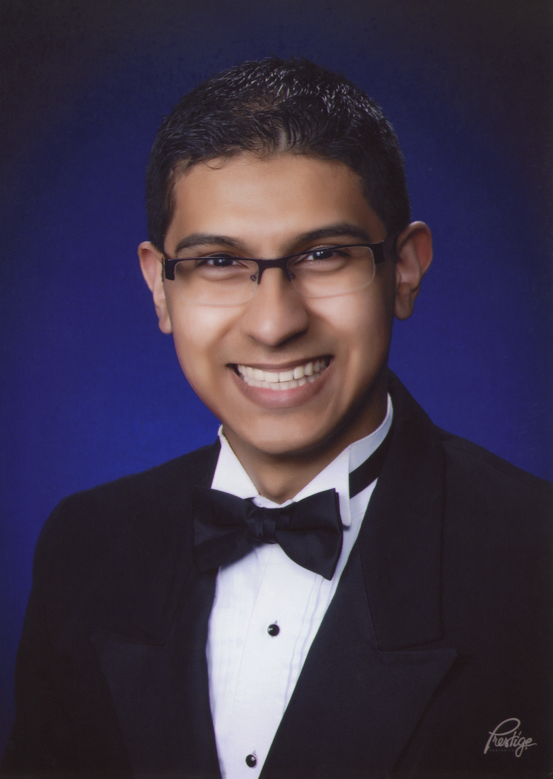 Kauri Allala wearing glasses, a tuxedo, and a bow tie, smiling against a dark blue background.
