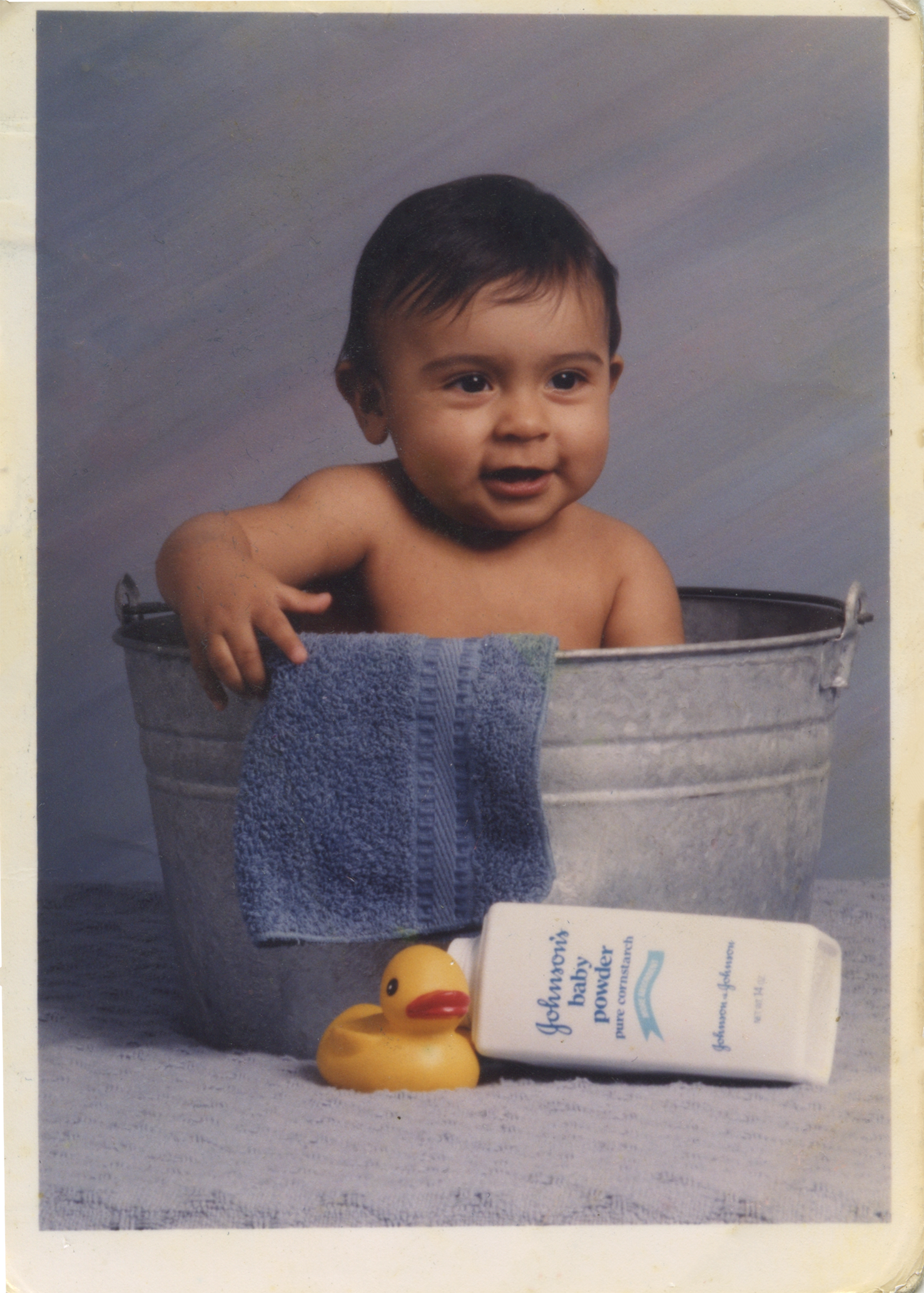 Kauri Allala, a baby boy with dark hair sitting in a metal tub, smiling, with a blue towel draped over the edge, set against a blue background; rubber duck and Johnson baby powder box on a surface in front.