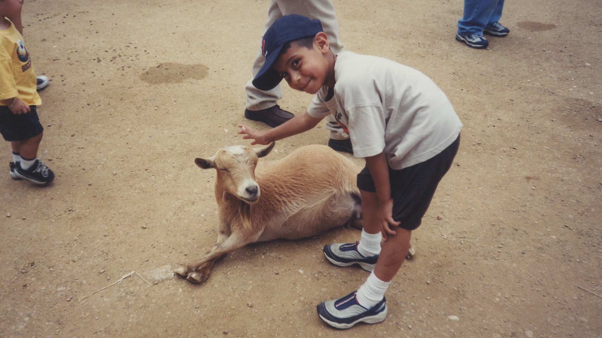 Kauri Allala wearing a baseball cap and shorts is bending over with a small goat laying on the ground next to him, with other children around on a dirt surface.