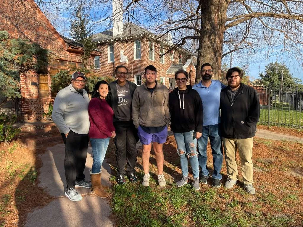 Group of seven Kauri's Lucky Star Foundation board members standing outdoors on a sidewalk in front of a large tree and brick houses, smiling for a photo on a sunny day.