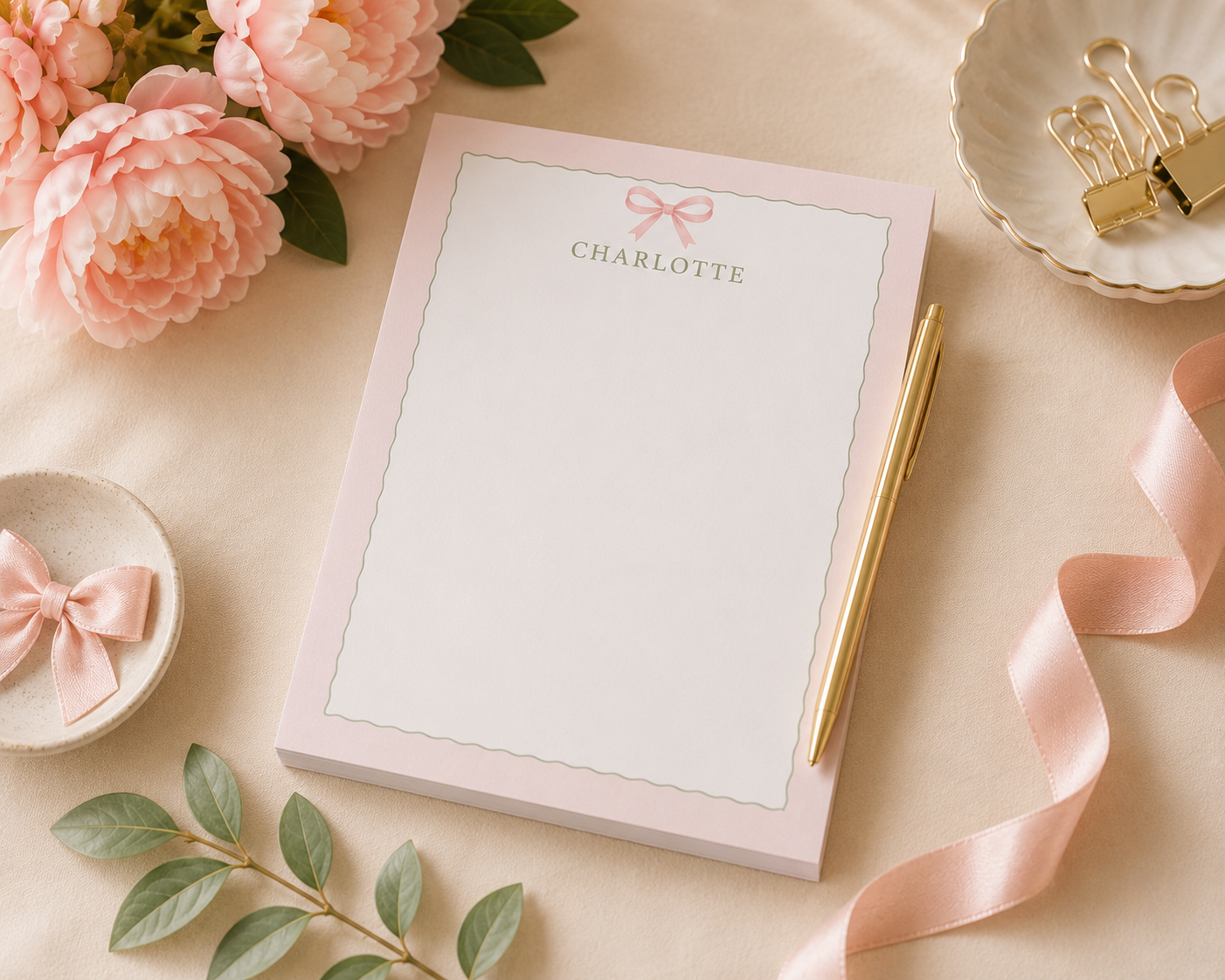 A pink notepad with personalized name at the top, decorative pink bow, and green border, surrounded by pink flowers, gold pen, pink ribbon, small dish with pink bow, and a decorative bowl with gold paper clips on a beige surface.