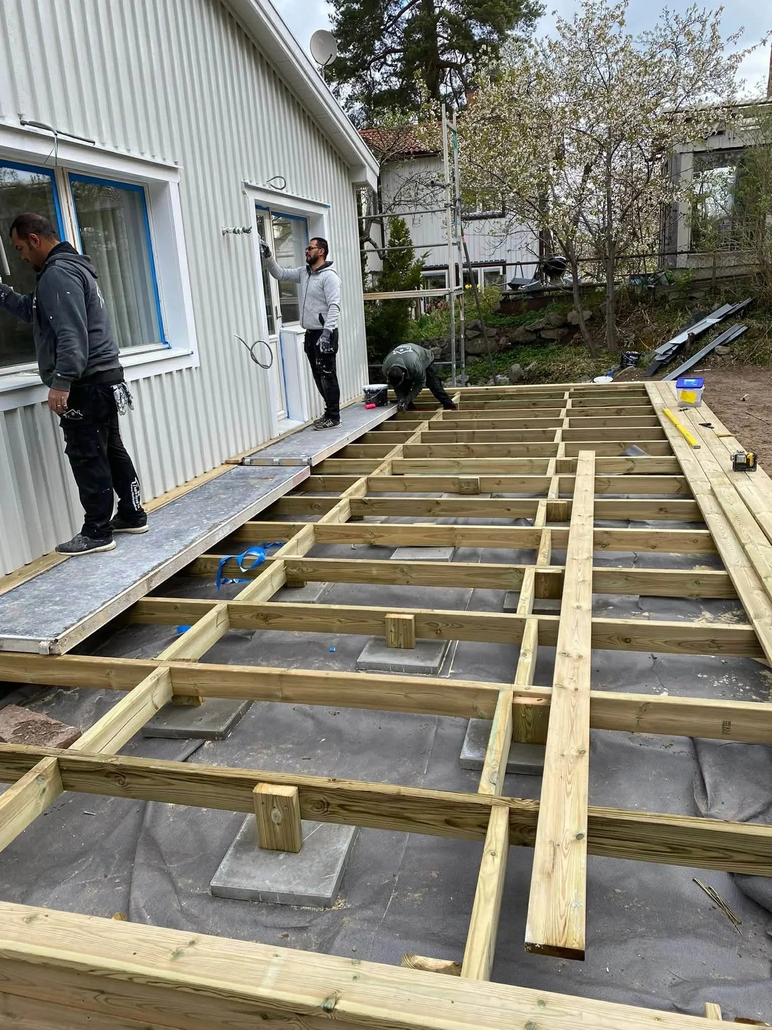 Byggarbetare som arbetar på en träkonstruktion utanför ett hus, med några träd och grönska i bakgrunden.