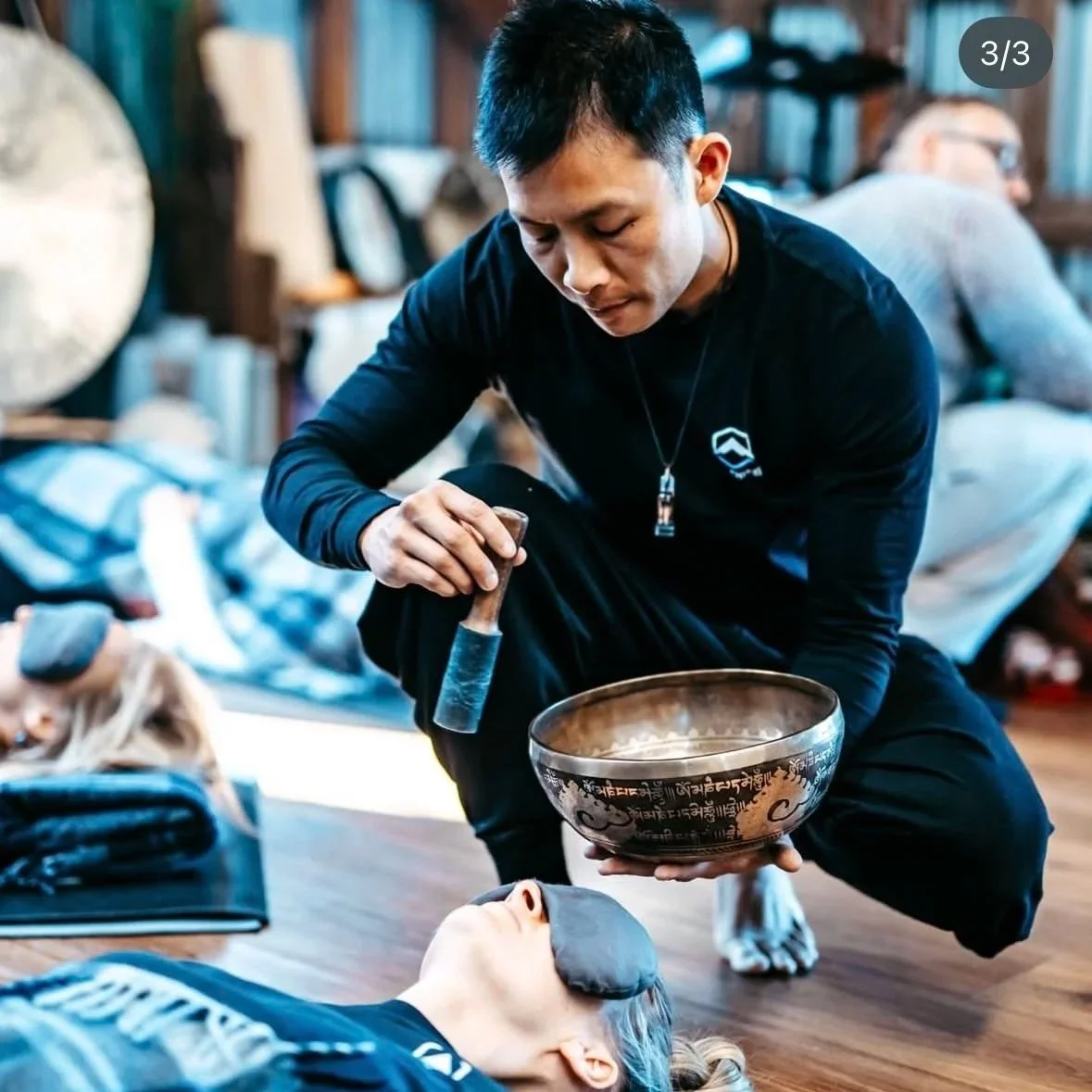 A person with short black hair, dressed in black, is kneeling next to a person lying on the floor with a black patch over their eye. The kneeling individual is holding a singing bowl and a mallet, appearing to perform a sound healing or meditation se