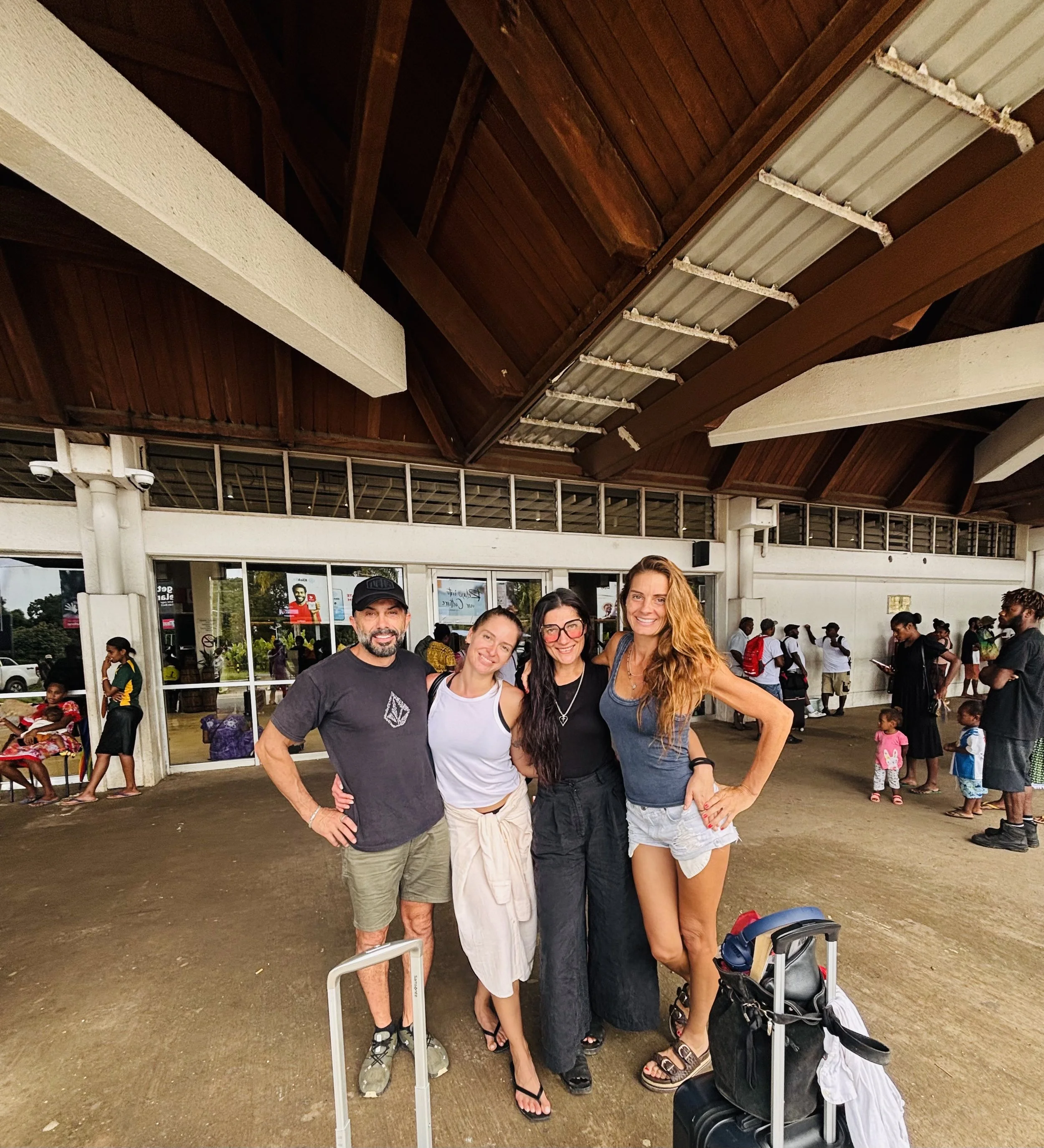Group of four friends smiling and posing at an airport terminal, with luggage and other travelers in the background.