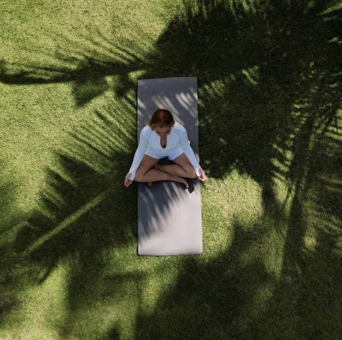 Overhead view of a woman sitting cross-legged on a yoga mat on grass, surrounded by shadows of palm trees.