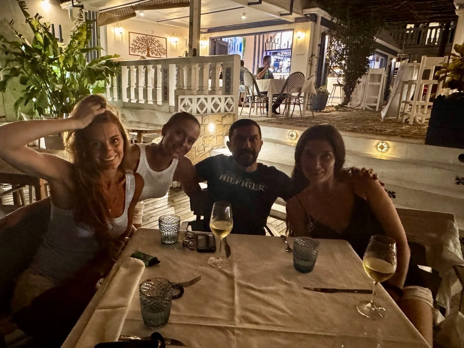 Four people sitting around a table at an outdoor restaurant during the evening. The table has wine glasses, candles, and utensils. There are plants and a lit building with patio furniture in the background.