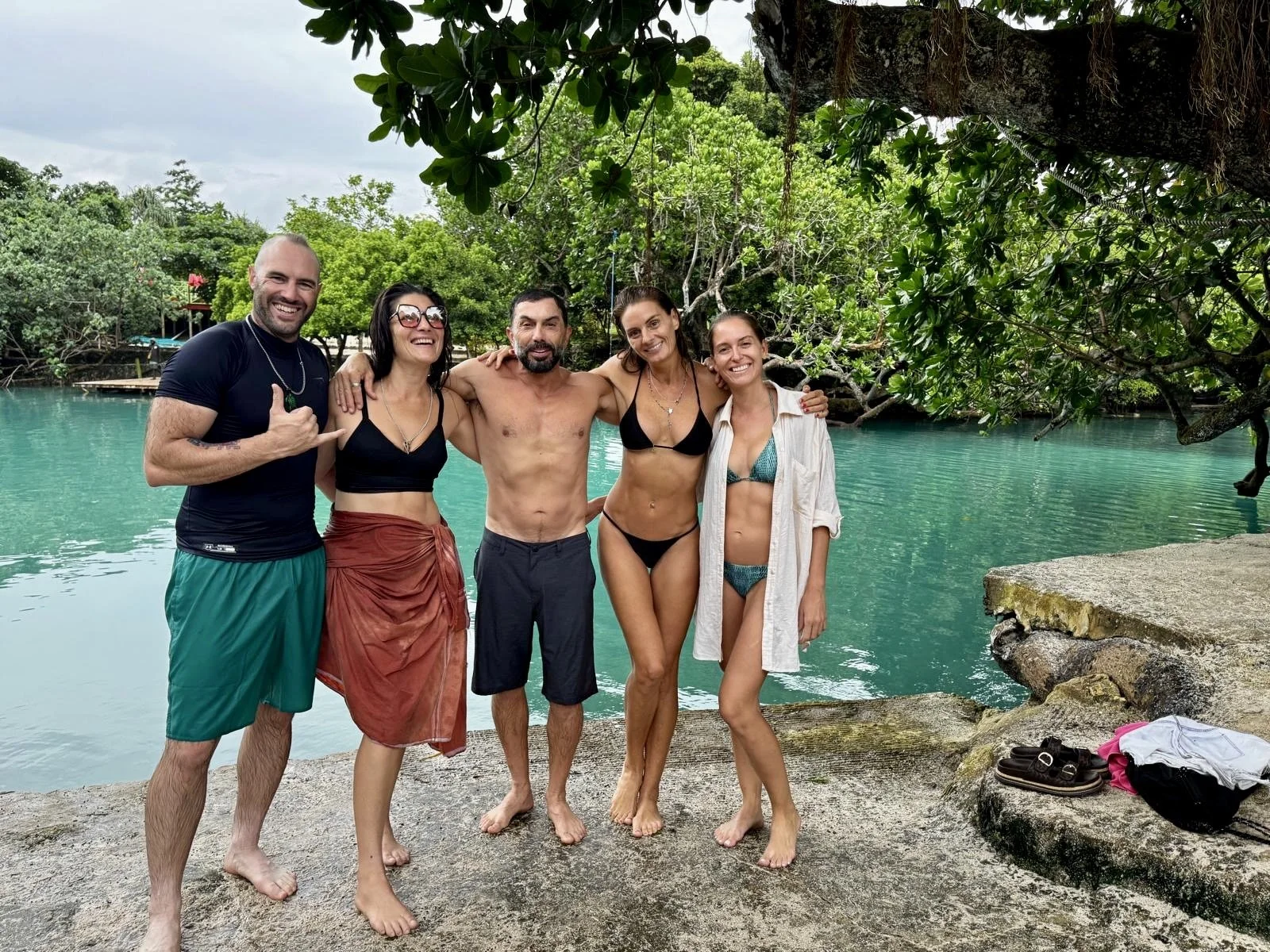 Five friends in swimsuits and casual clothing standing on rocks near a turquoise river, smiling and posing with arms around each other, in a lush, green outdoor setting.