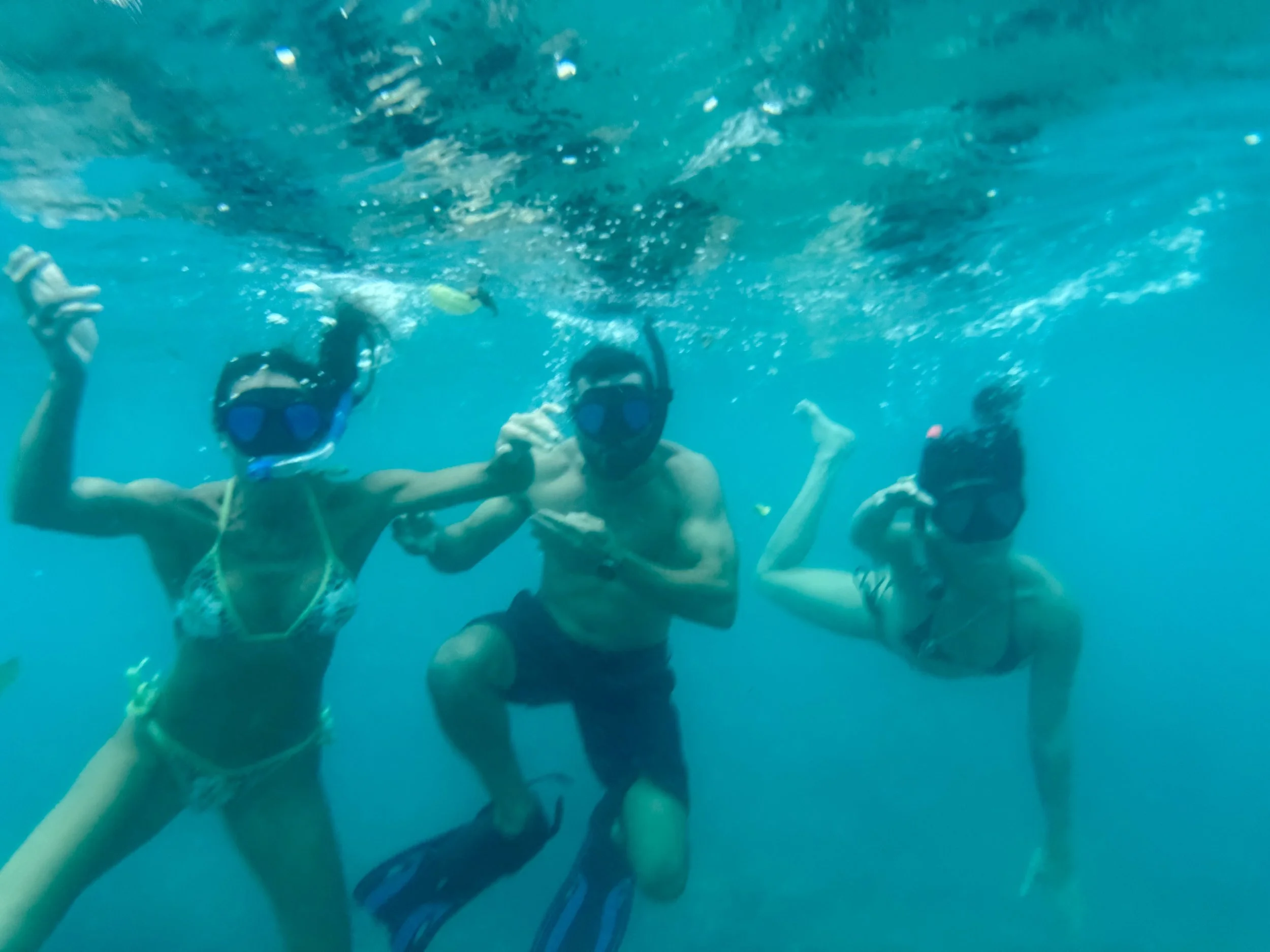 Three people in bikinis and swim masks underwater, holding hands and posing for a photo.