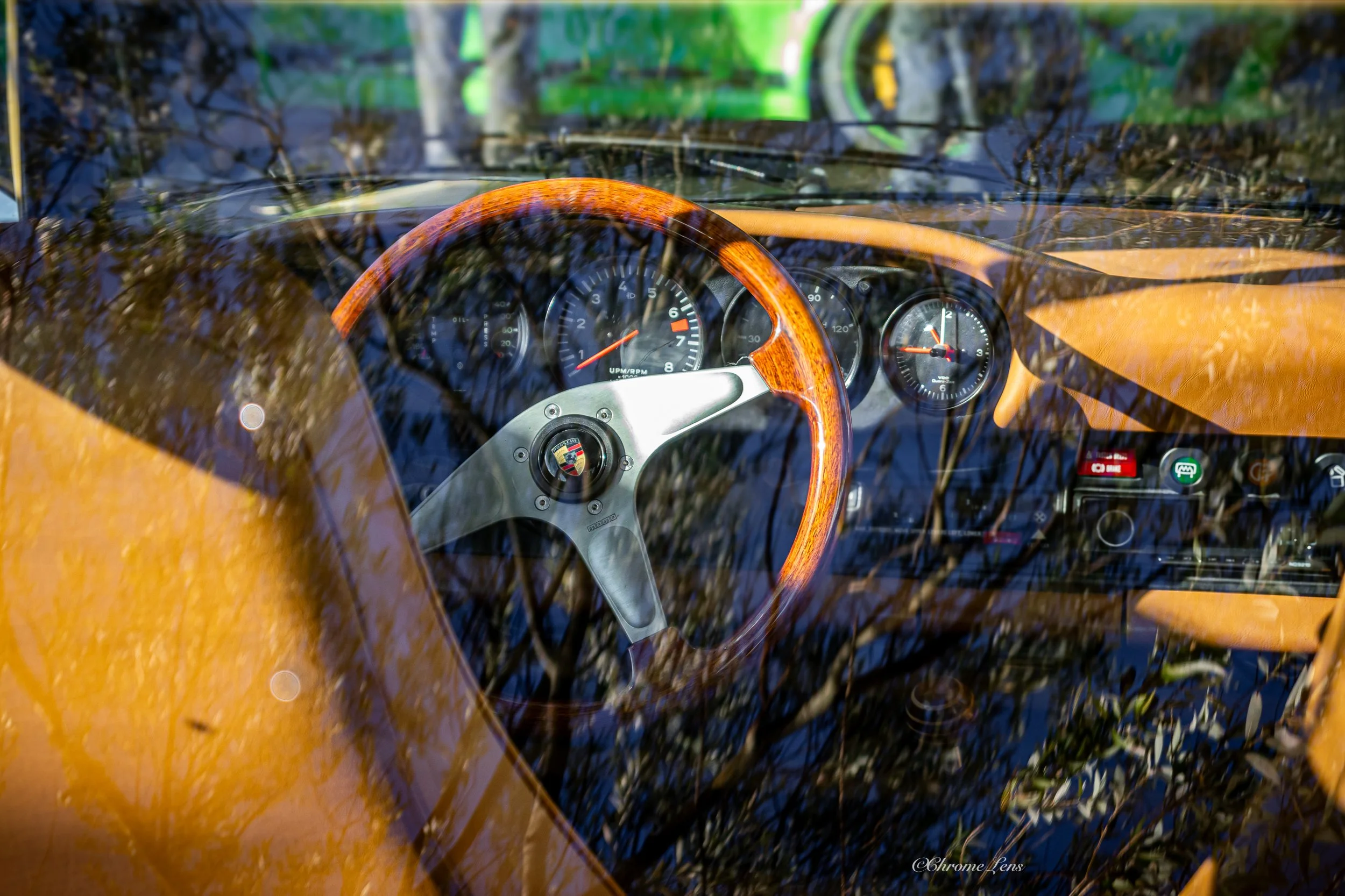 View of a Porsche 911 S  interior through the windshield, featuring a wooden steering wheel, vintage dashboard gauges, and a reflection of trees.