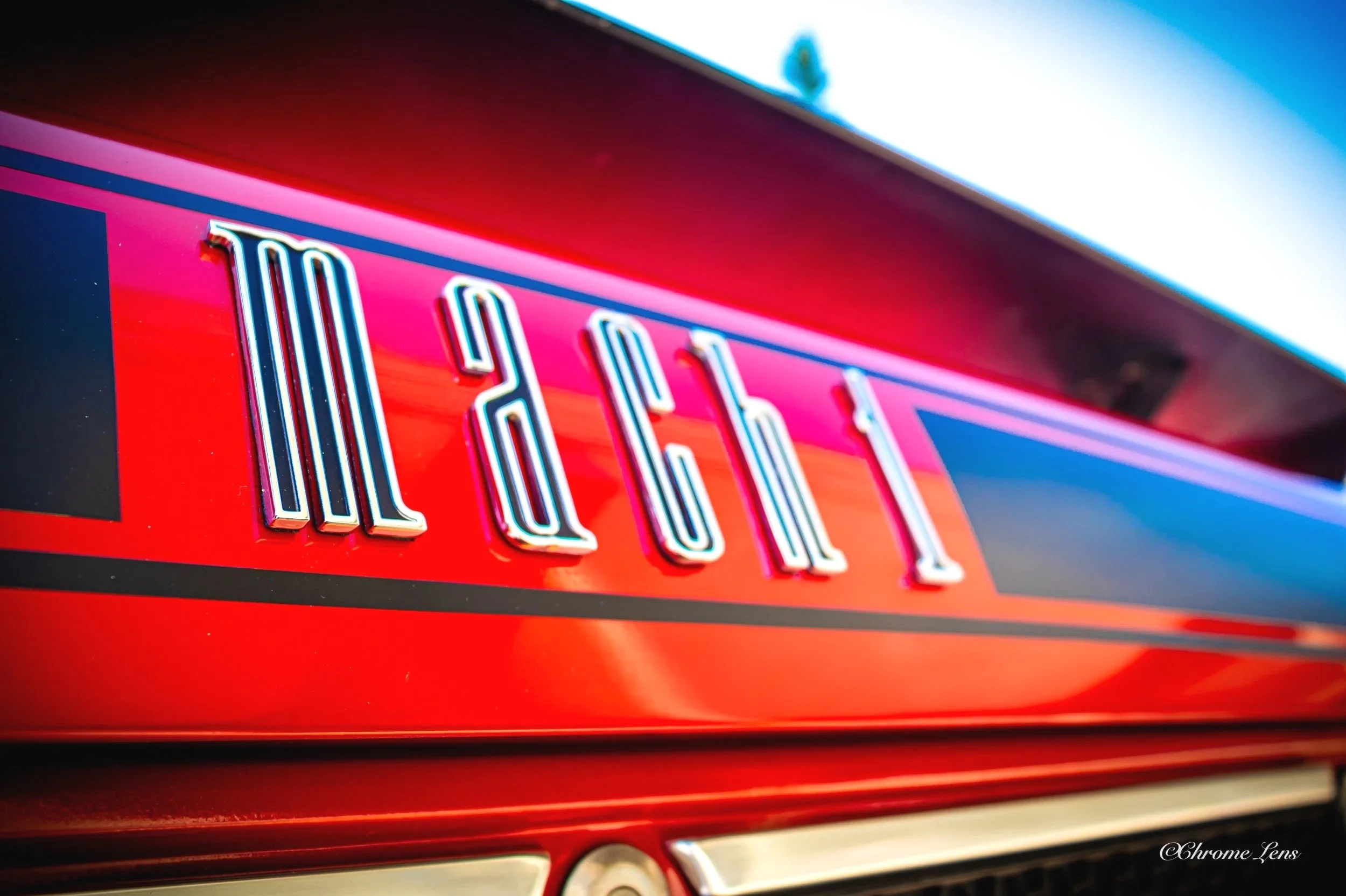 Close-up of a red Mustang Mach 1 with chrome letters spelling 'Mach 1r' on the back, reflecting the sunlight.