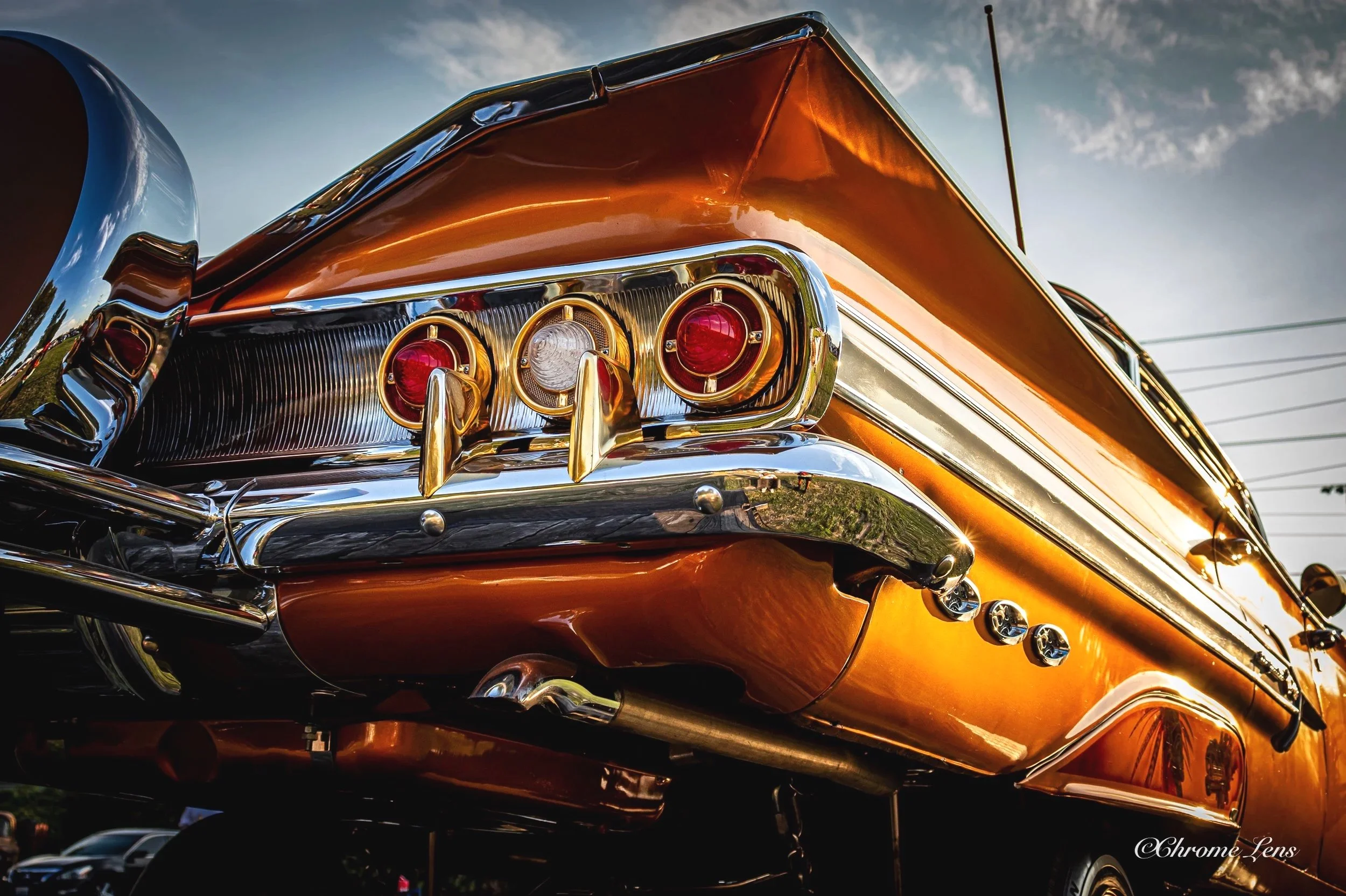 Close-up of the rear end of a vintage orange and chrome car with tail lights and decorative elements, under a cloudy sky.