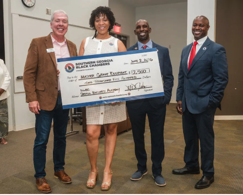 Four people standing indoors, holding a large check from Southern Georgia Black Chambers, with a woman in the center holding the check, all dressed in business attire, smiling at the camera.