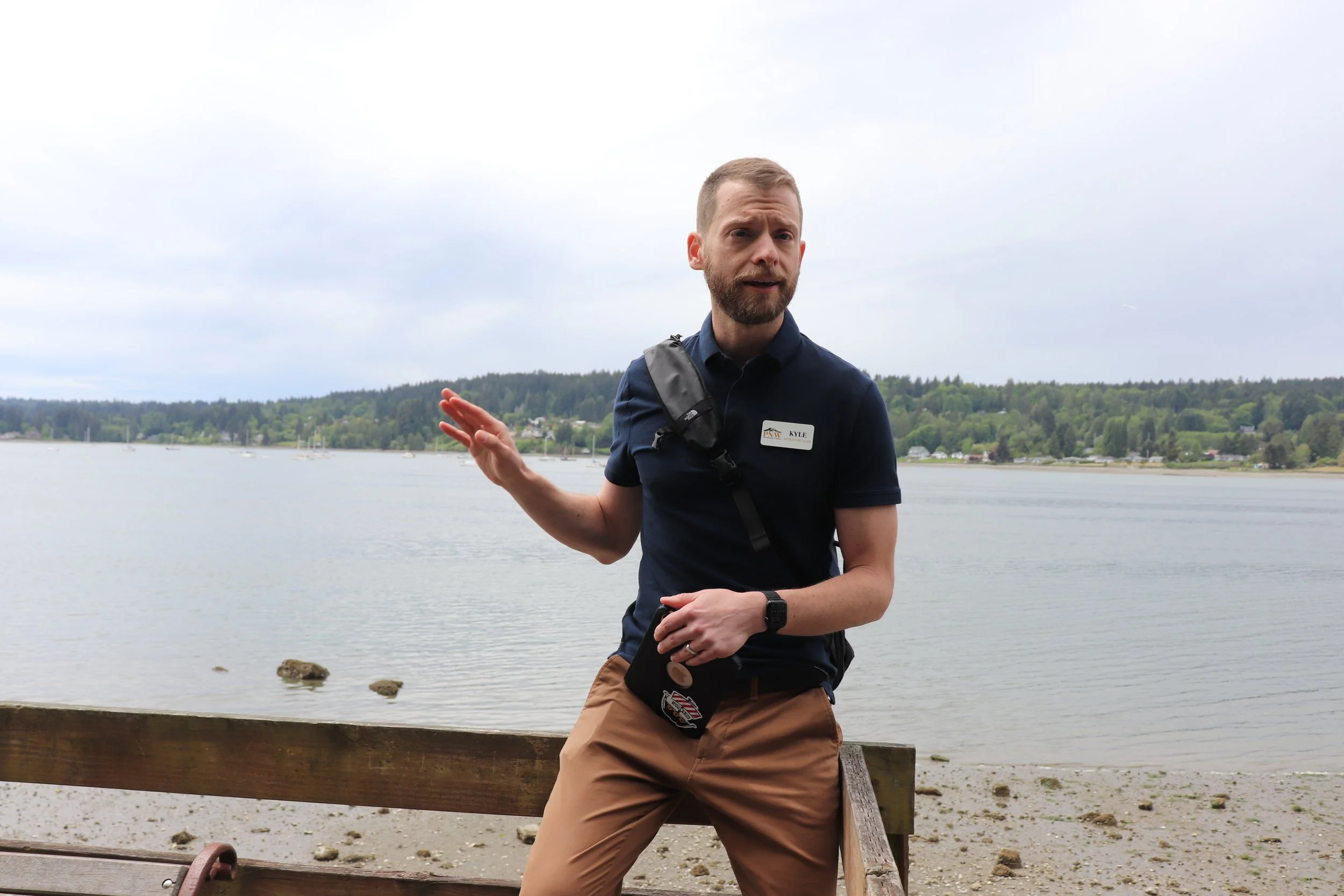 A man with light skin, short hair, and a beard standing outdoors near a body of water, wearing a navy blue polo shirt with a name tag and a shoulder bag.