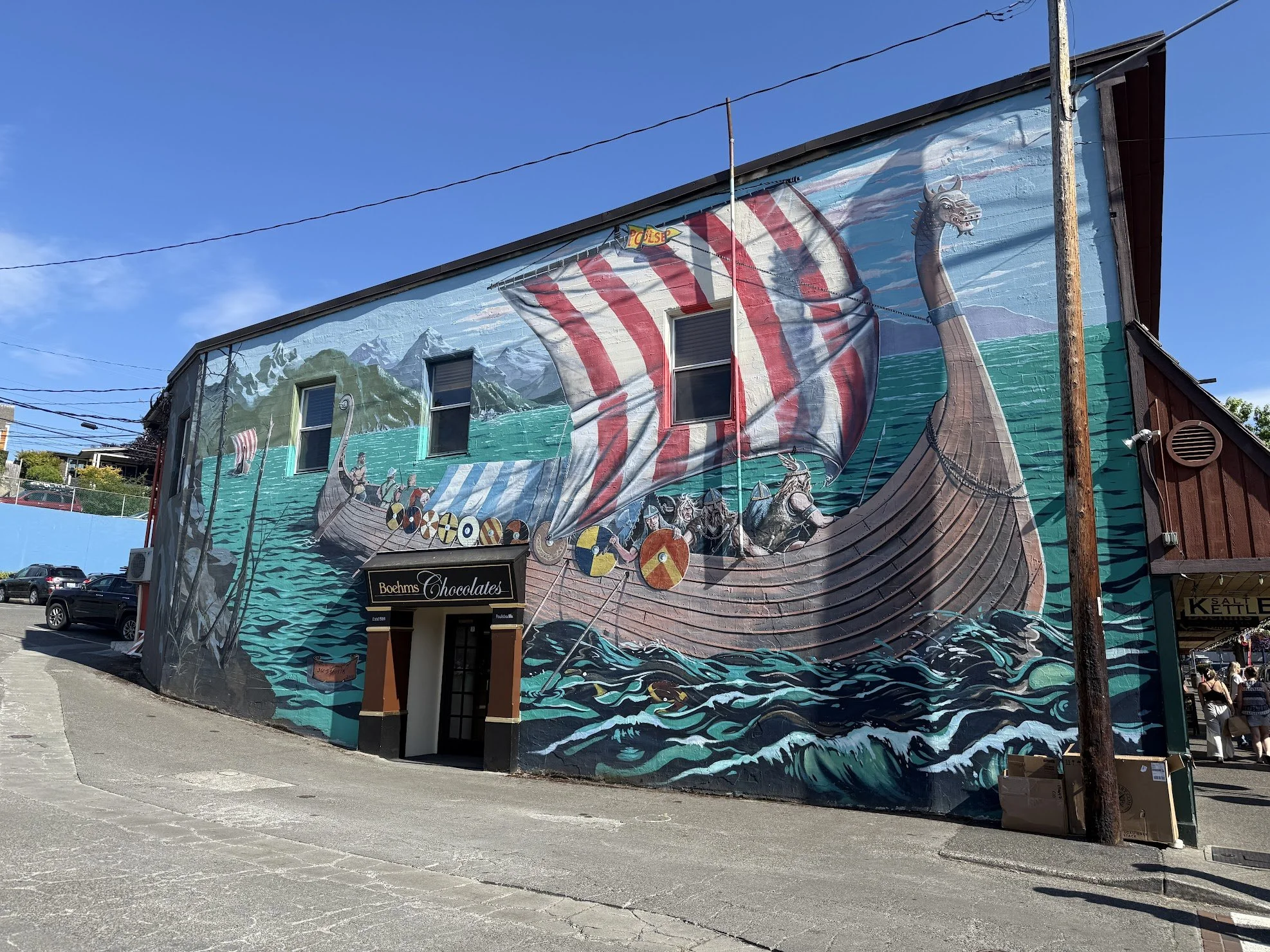 Colorful mural on the side of a building depicting a Viking ship with a red and white striped sail, Vikings, and water waves beneath, with a mountain background.