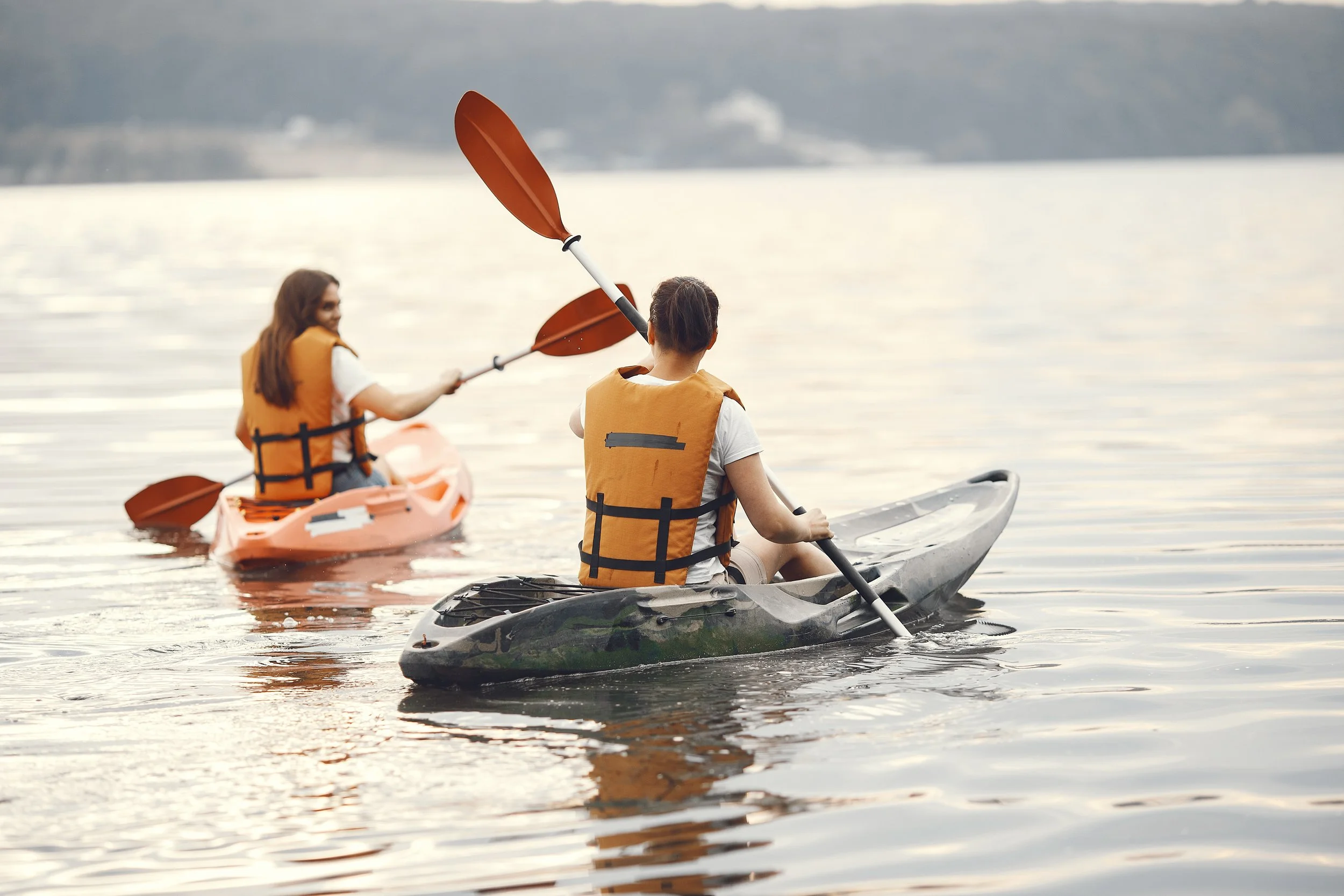 Two women kayaking on a calm lake, wearing orange life jackets, with a distant tree-lined shoreline in the background during sunset.