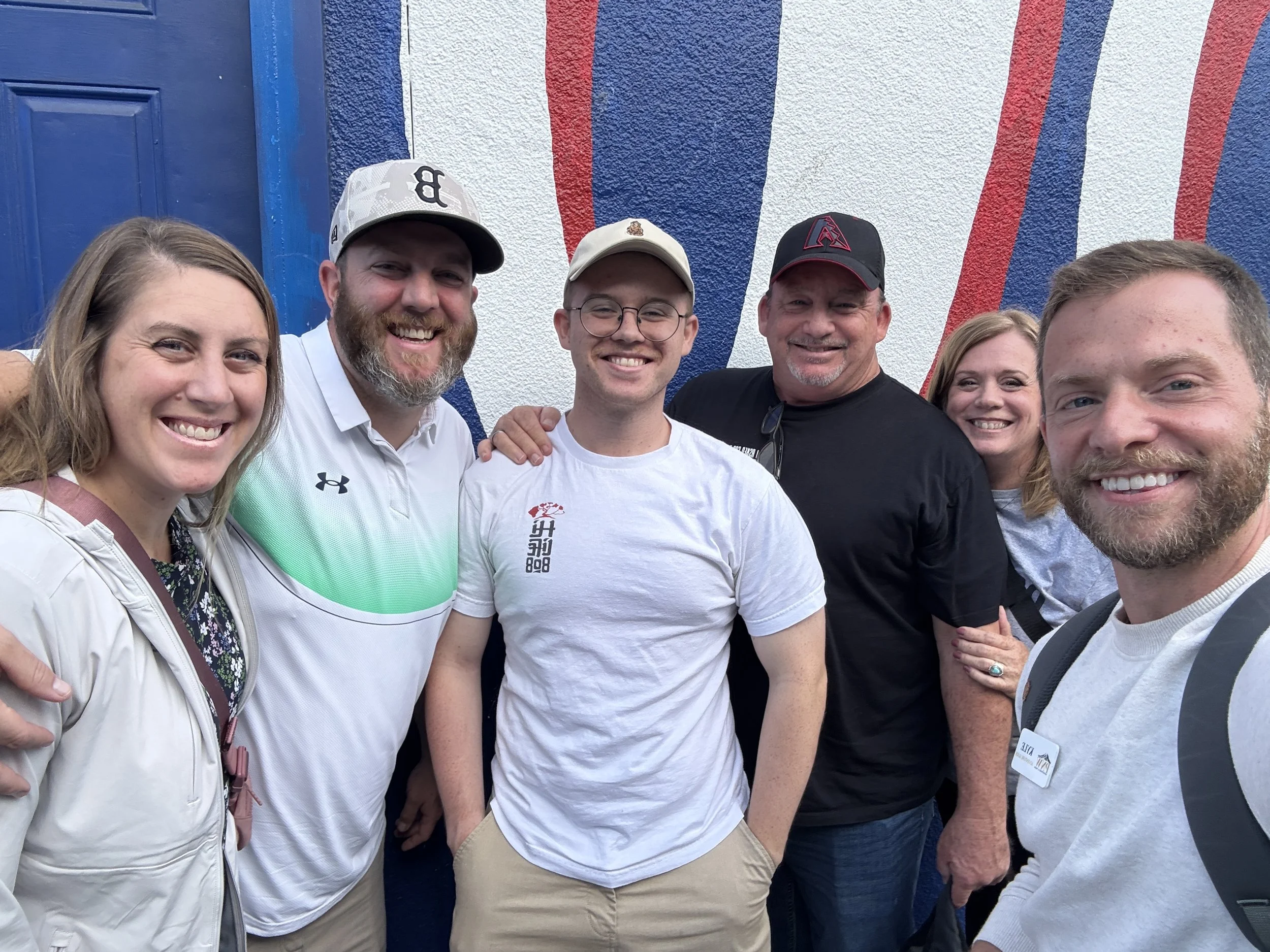 A group of six people standing close together outdoors, smiling for a selfie, with a colorful mural painting in the background.