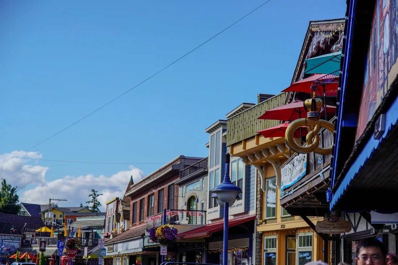 A busy street with colorful storefronts, hanging umbrellas, and a blue sky with clouds.