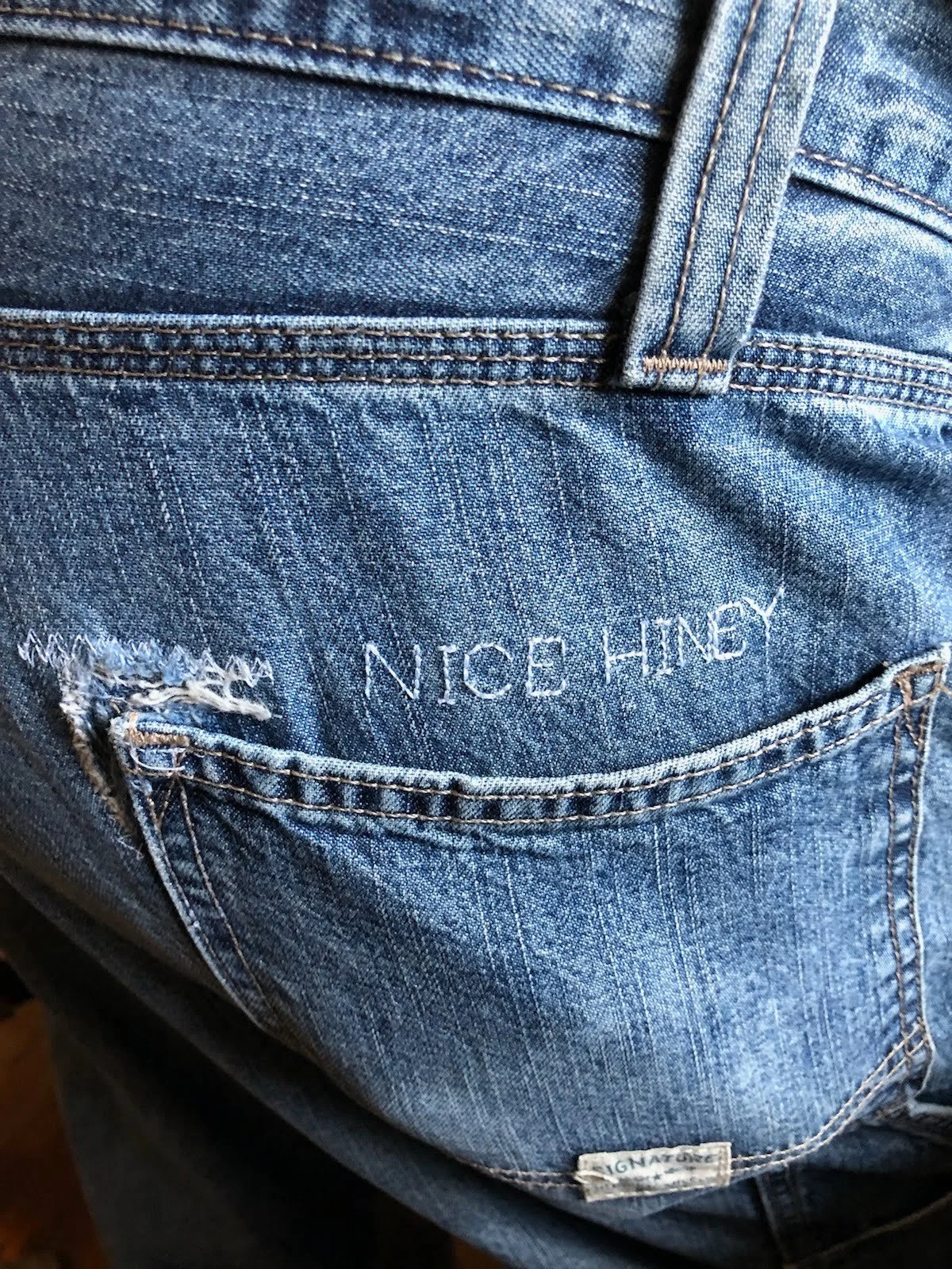 Close-up of blue denim jeans with the embroidered name "Nice Hiney" on the back pocket.