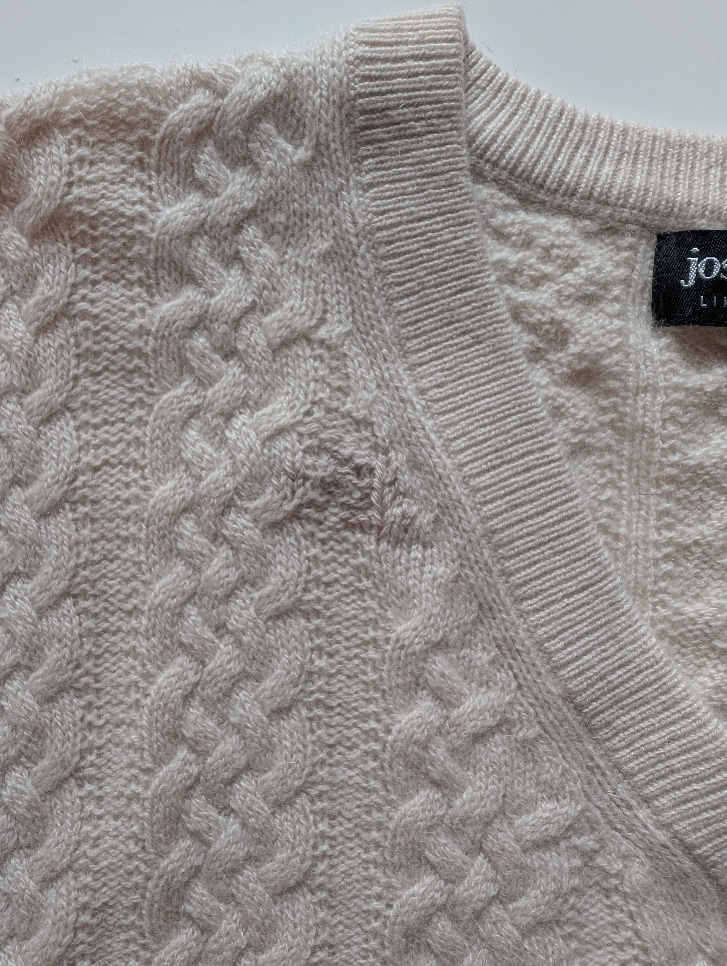 Close-up of a beige cable-knit sweater with a ribbed neckline and a black brand tag.
