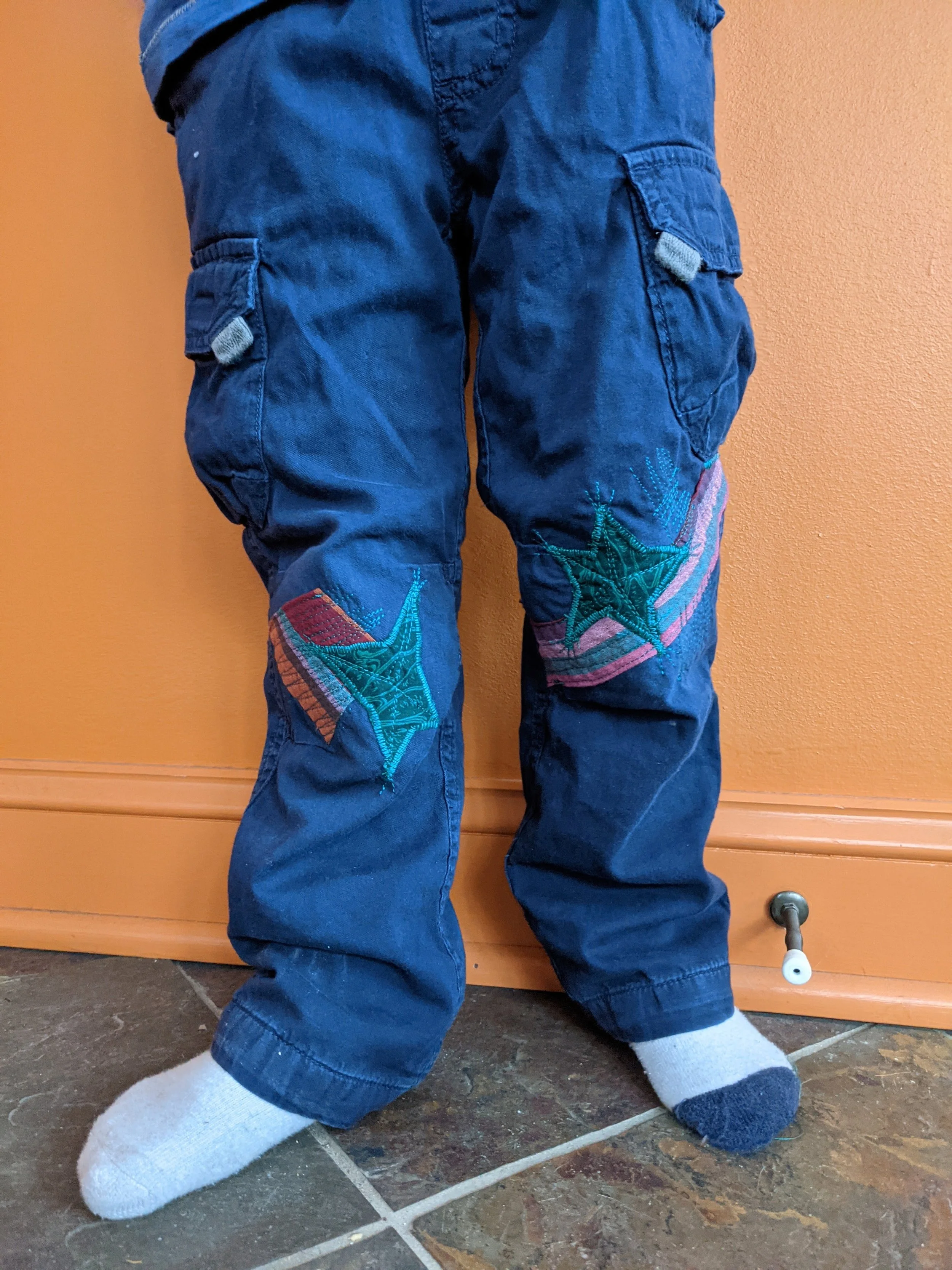 Kids wearing dark blue cargo pants with embroidered star patches on knees, standing on a tiled floor in front of an orange wall.
