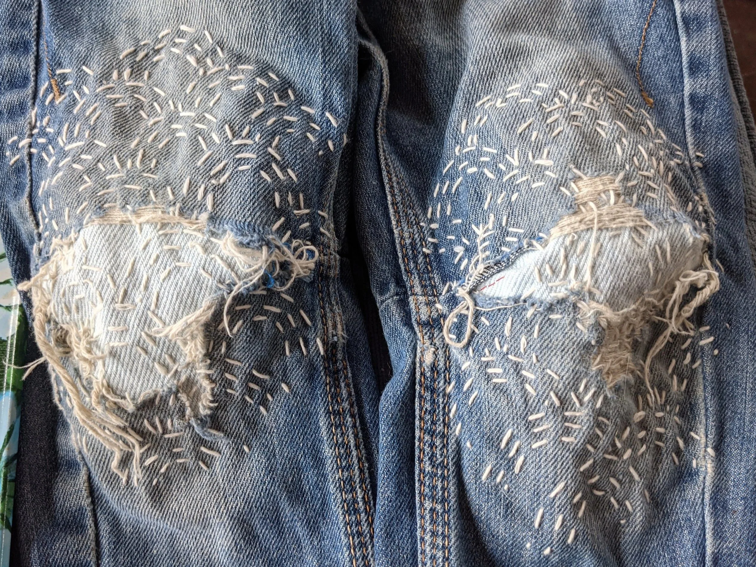 Pair of blue denim jeans with large torn patches on the knees, stitched with white thread in decorative patterns