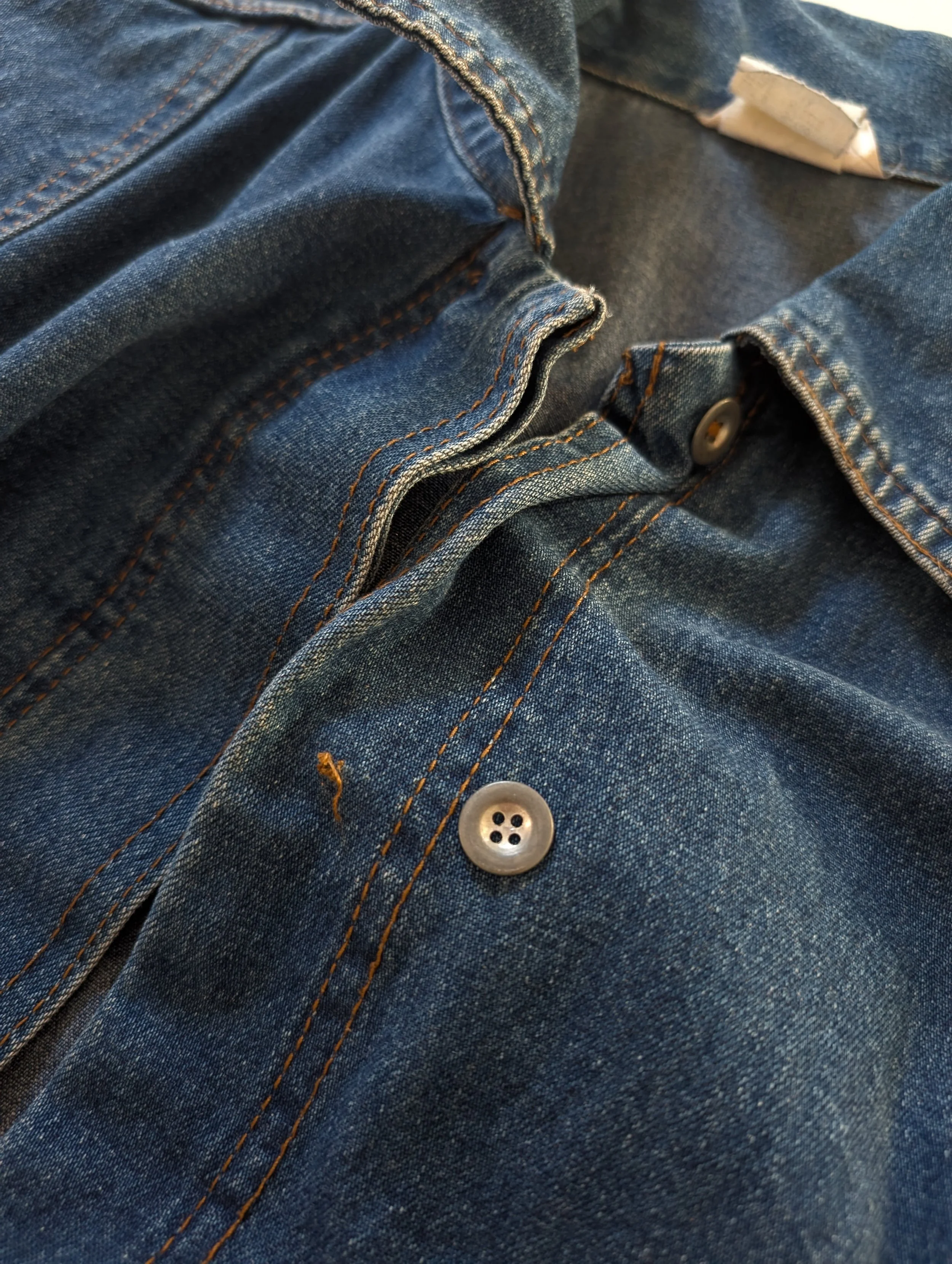 Close-up of a blue denim shirt with orange stitching and gray buttons, showing the fabric and button details.