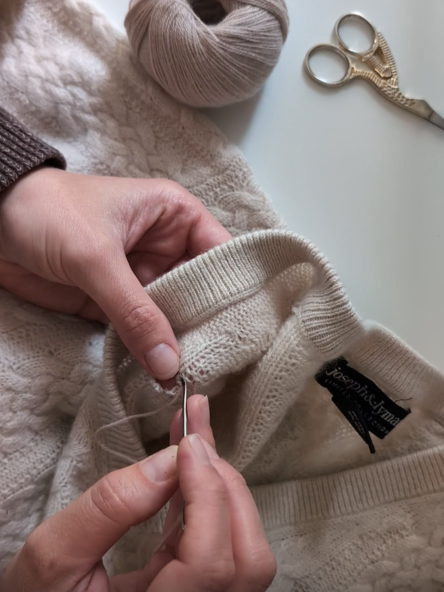 Person sewing a beige sweater with a needle, surrounded by yarn and scissors.