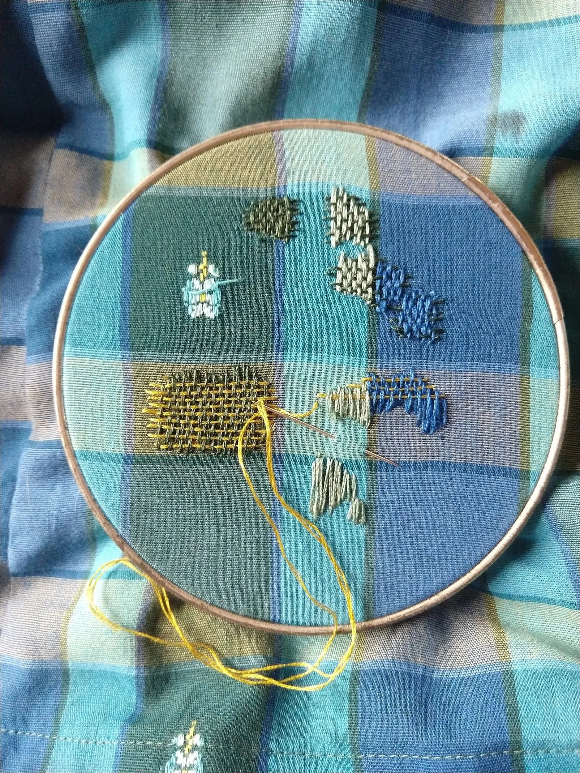 Embroidery hoop with partially completed cross-stitch design on plaid fabric, featuring various colored threads and stitches.