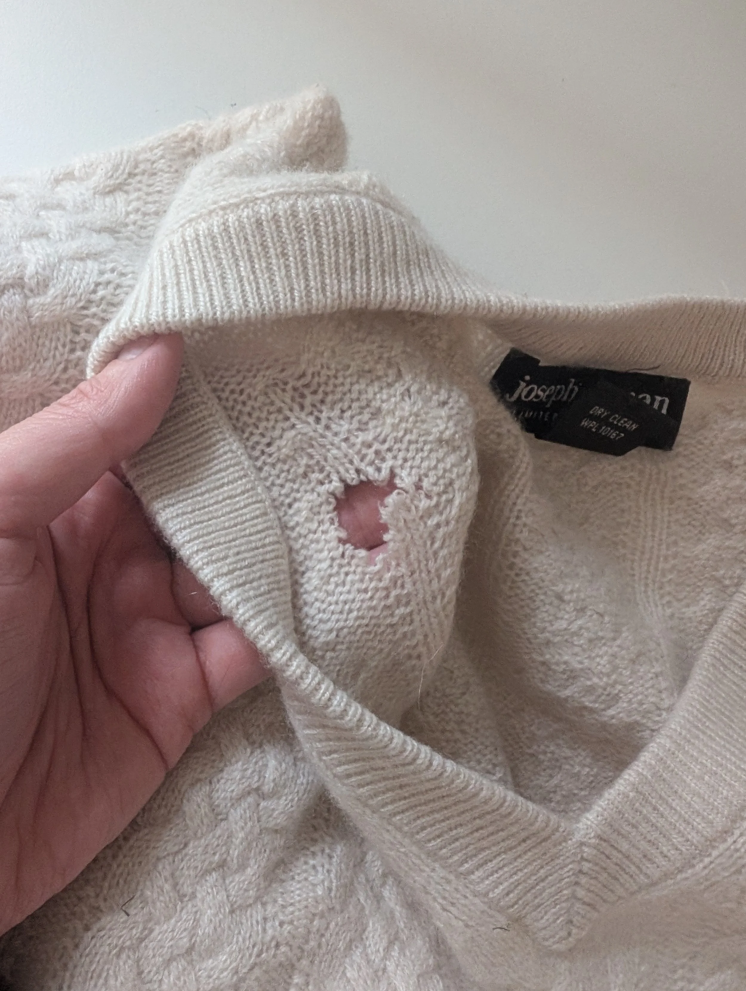 Close-up of a cream-colored knit clothing item with a tear, showing a hole in the fabric, held by a person's hand. The clothing has a black label that reads 'Joseph'.