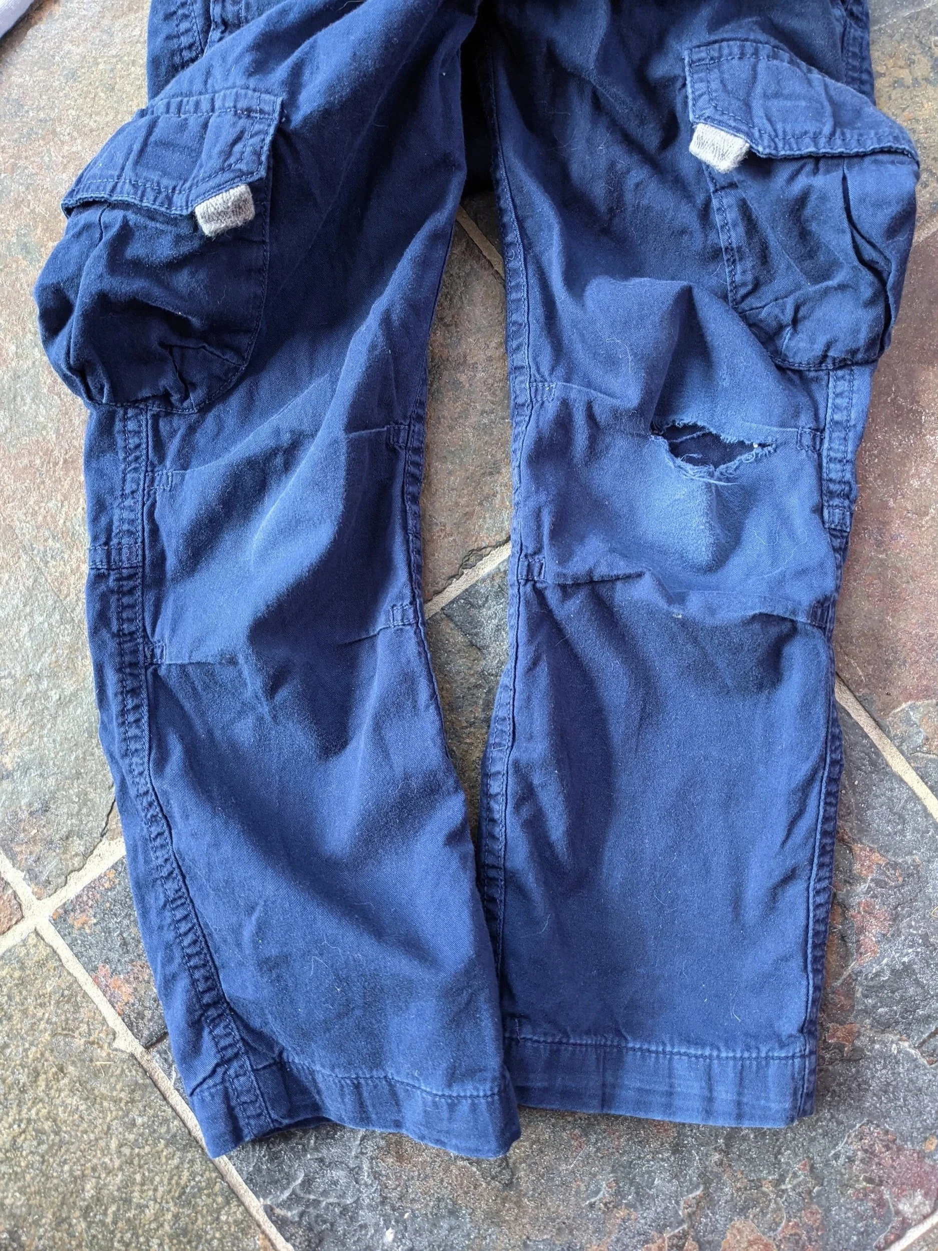 A pair of blue cargo pants with a tear on the right leg, placed on a tiled floor.