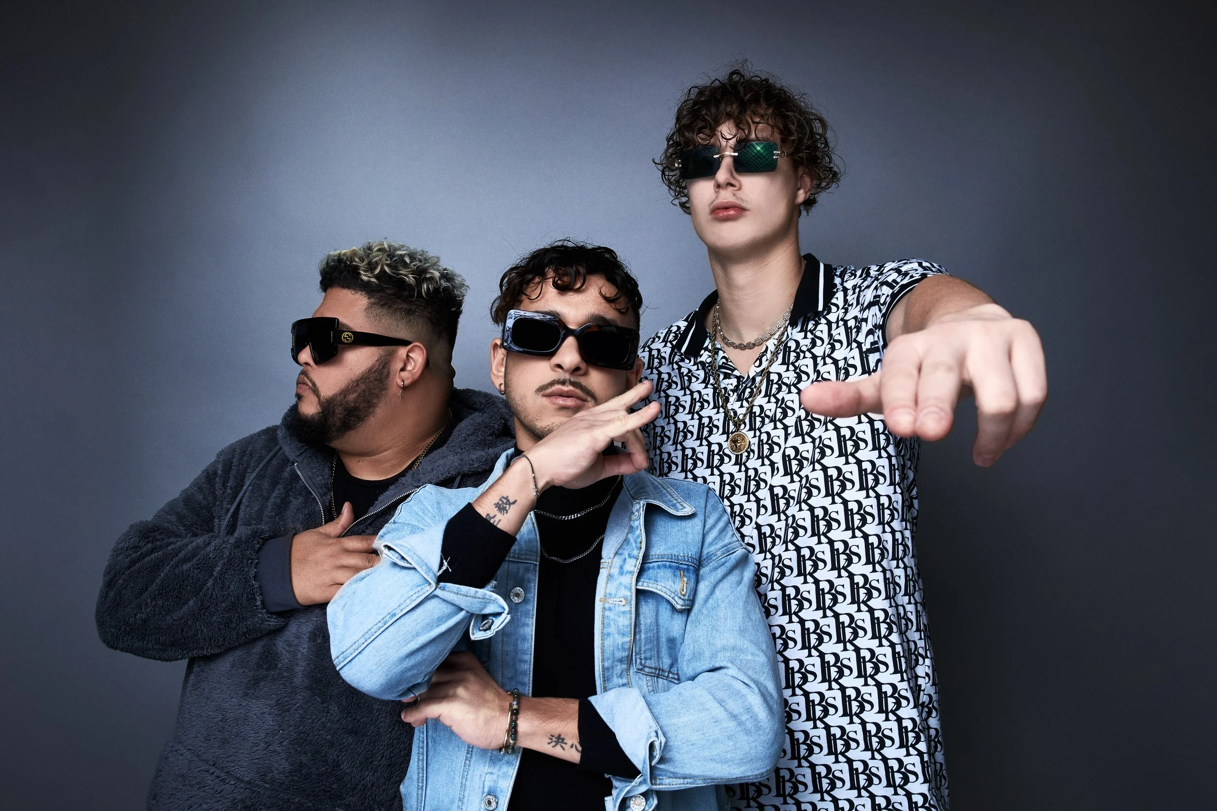 Three young men with curly hair and sunglasses posing confidently against a dark gray background, with one man pointing towards the camera.