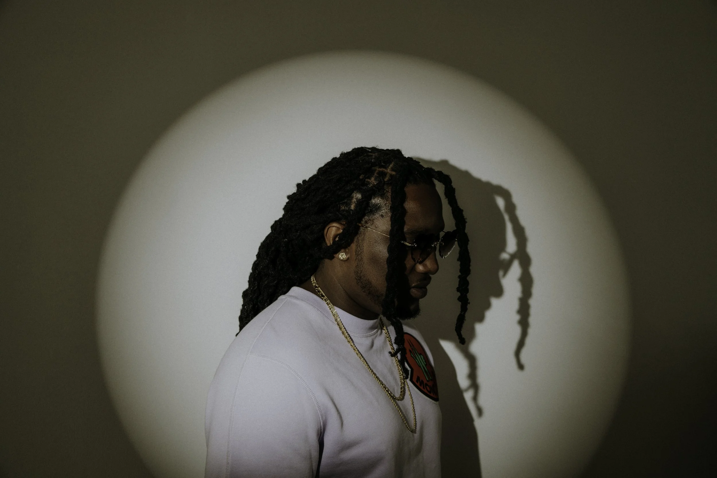 A young man with dreadlocks wearing sunglasses, gold chains, and a white shirt with a logo, standing in front of a plain background with a circular spotlight, casting a shadow of his profile on the wall.