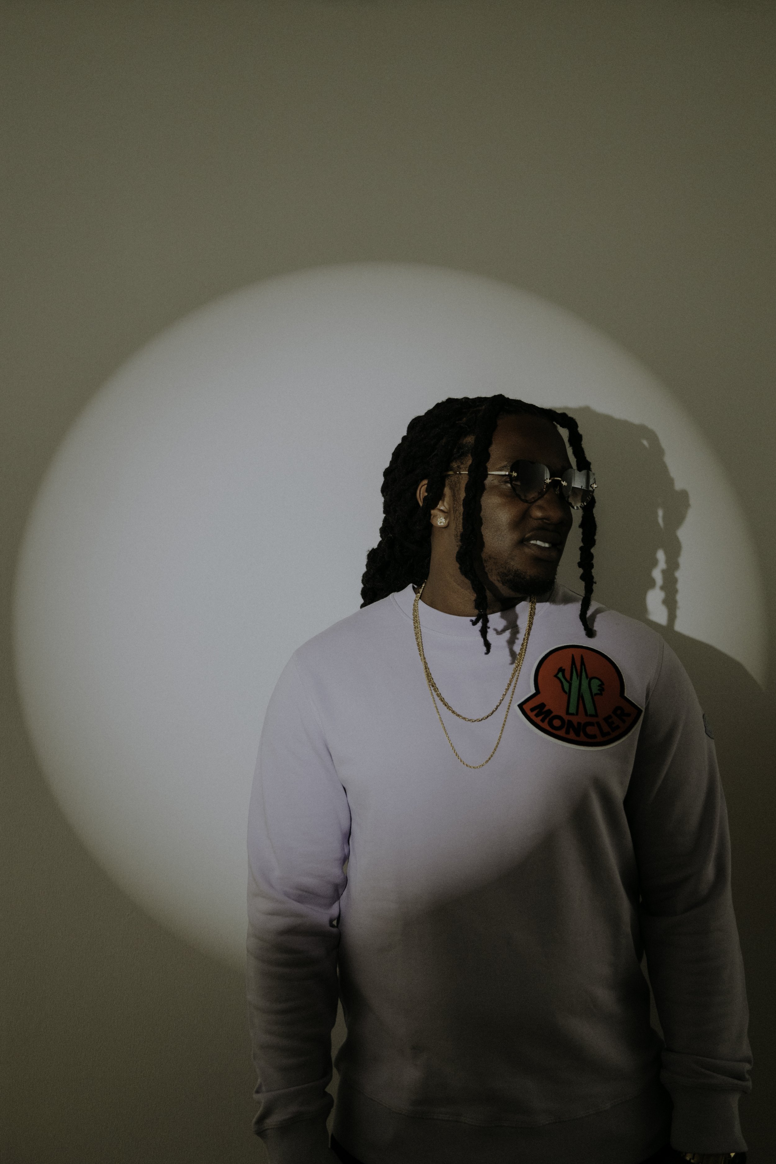 A man with long dreadlocks, glasses, and gold jewelry, wearing a white Moncler sweatshirt, stands in front of a wall with a circular light or projection behind him.