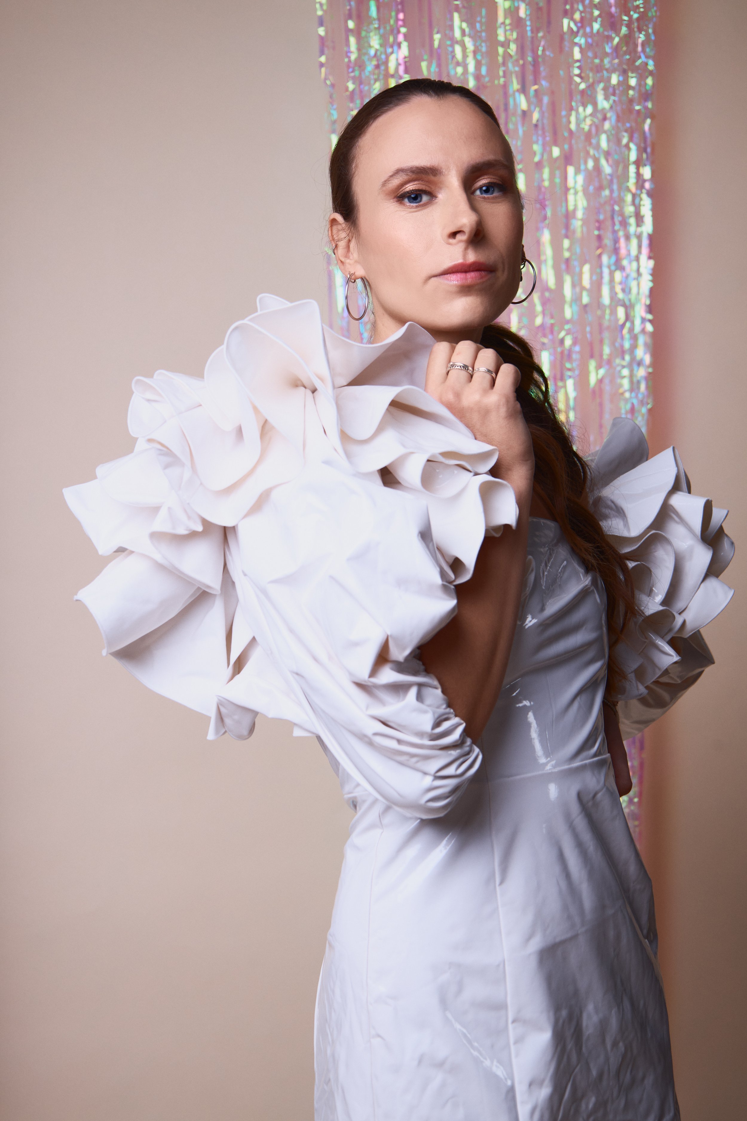 A woman with brown hair and blue eyes wearing a white dress with ruffled, sculptural sleeves, standing against a beige wall with iridescent decoration in the background.