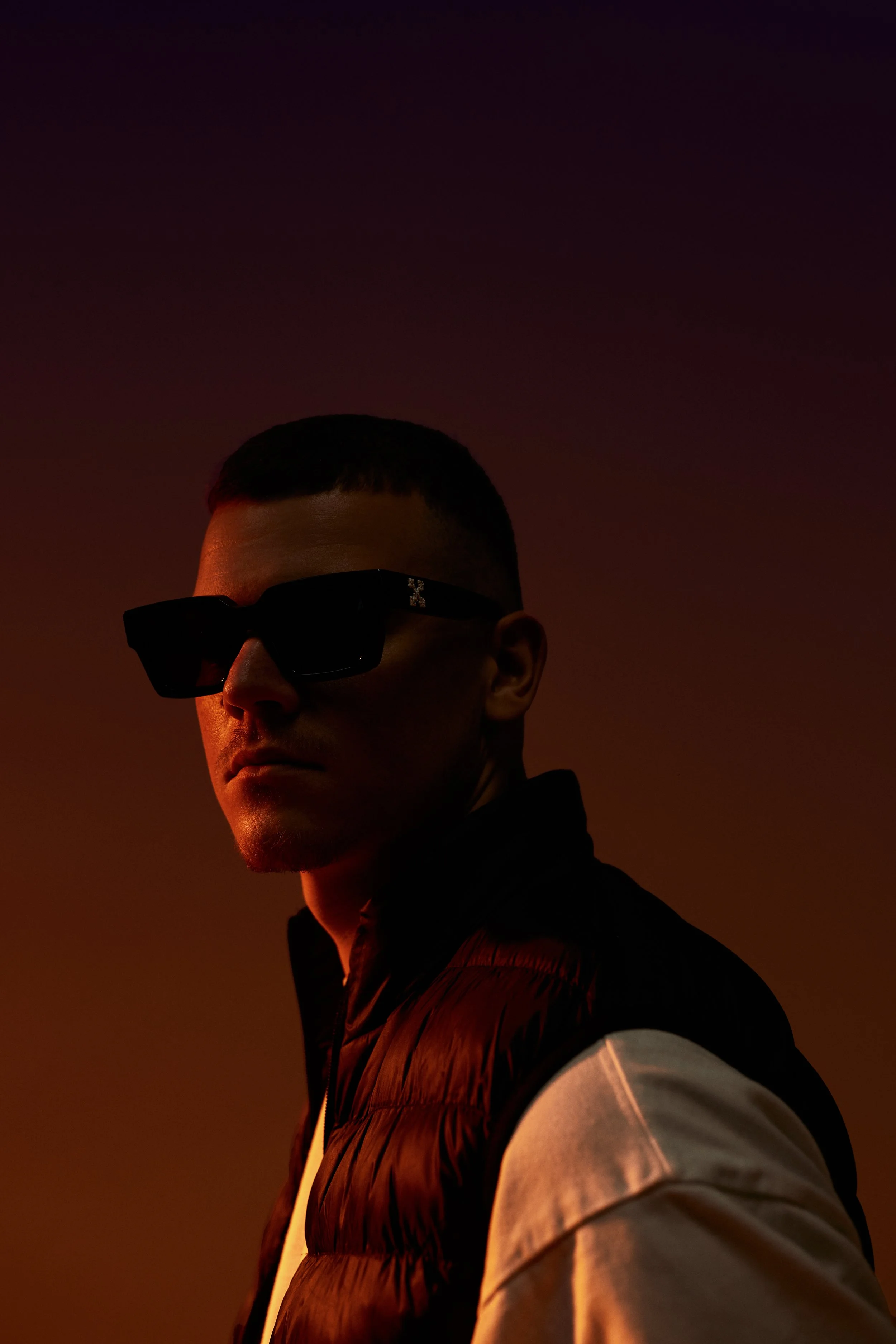 A man wearing dark sunglasses and a black vest against a gradient dark background.