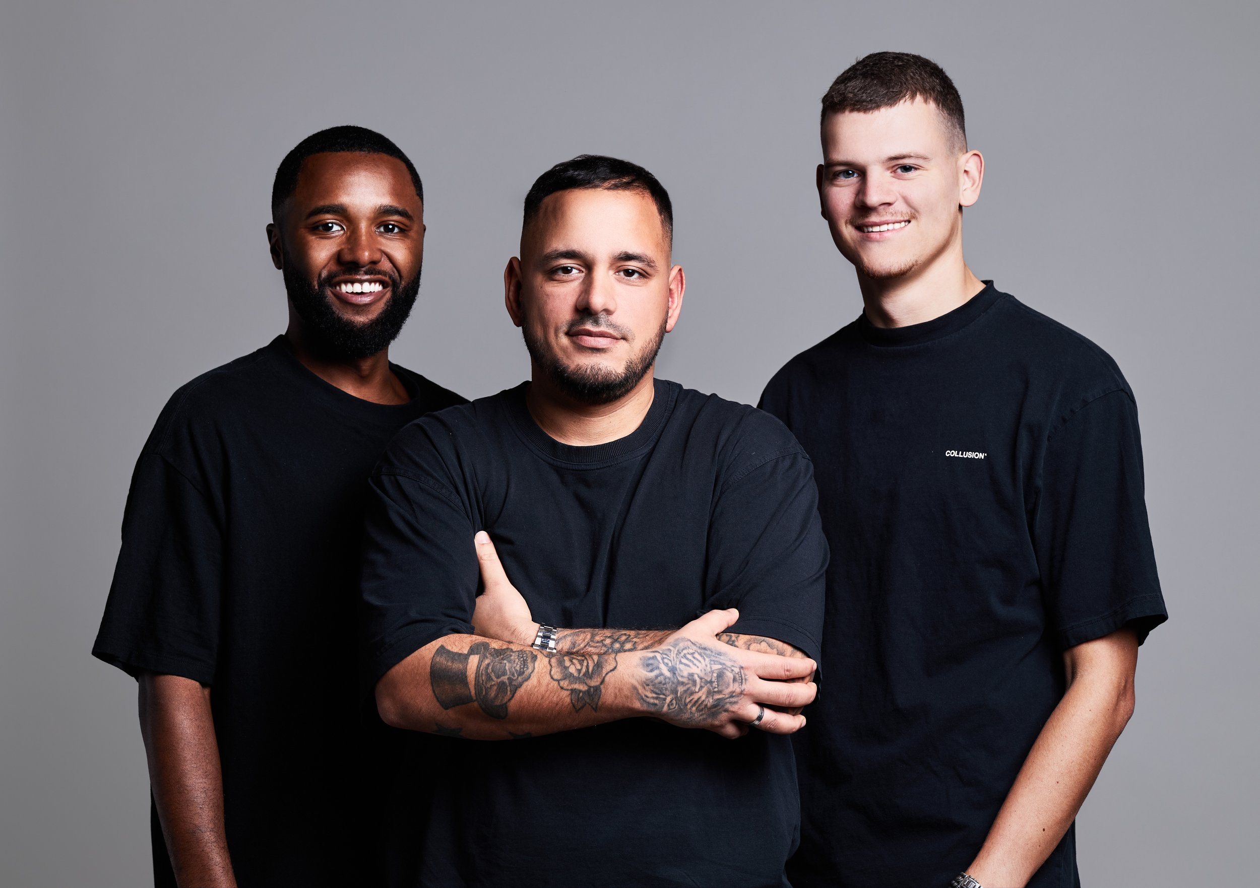 Three young men standing together against a gray background, all wearing black t-shirts, smiling and looking at the camera.