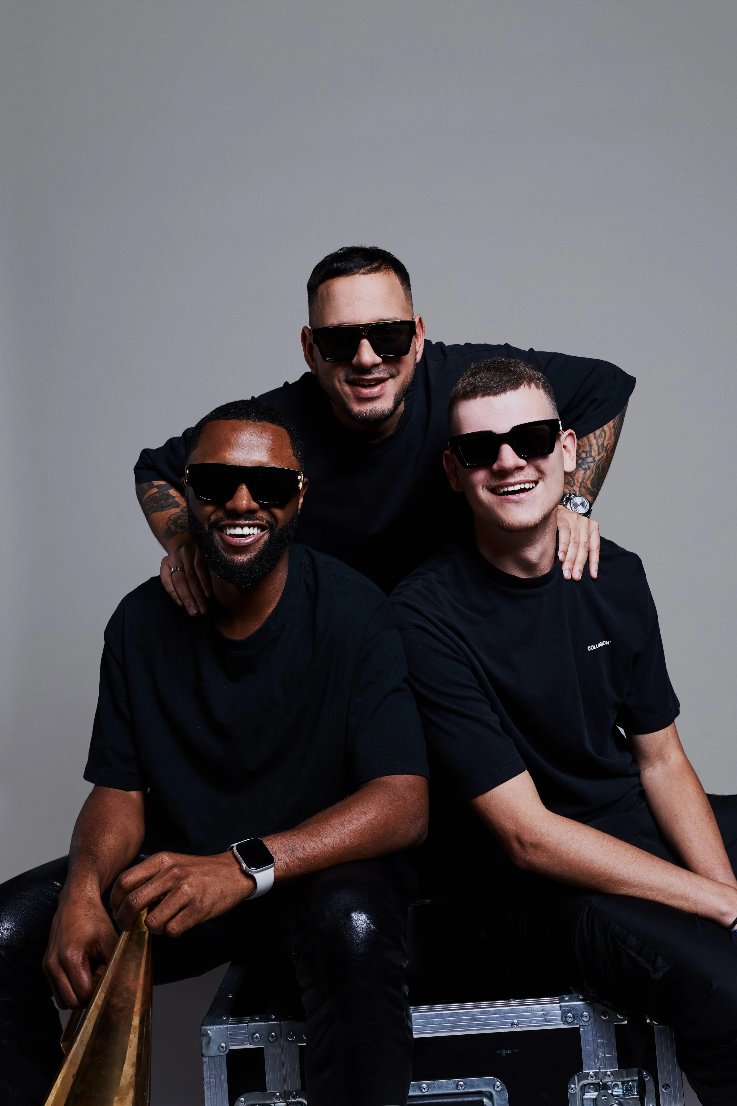 Three men wearing sunglasses, sitting and standing together against a plain gray background, smiling.