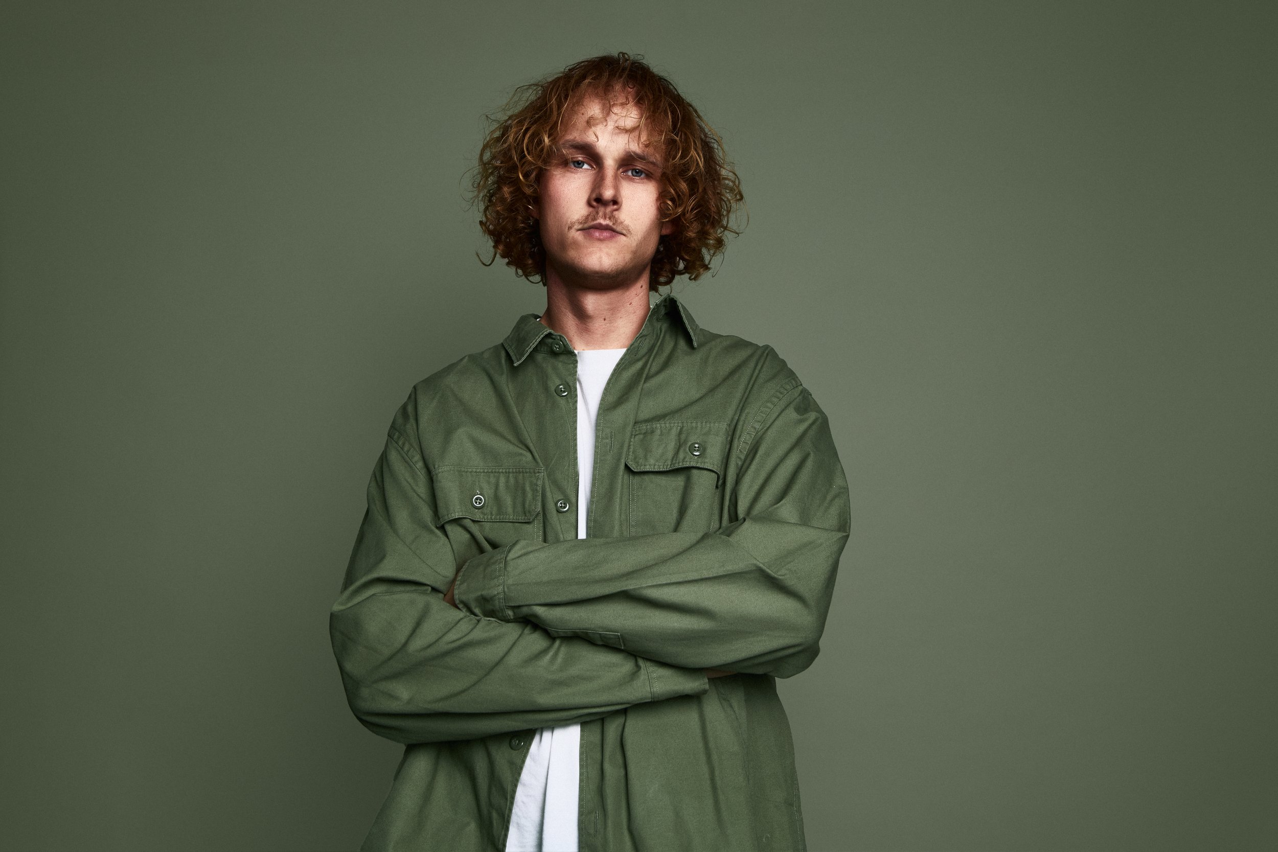 A young man with curly red hair and a serious expression, crossing his arms, wearing an oversized green shirt over a white t-shirt, standing against a plain green background.