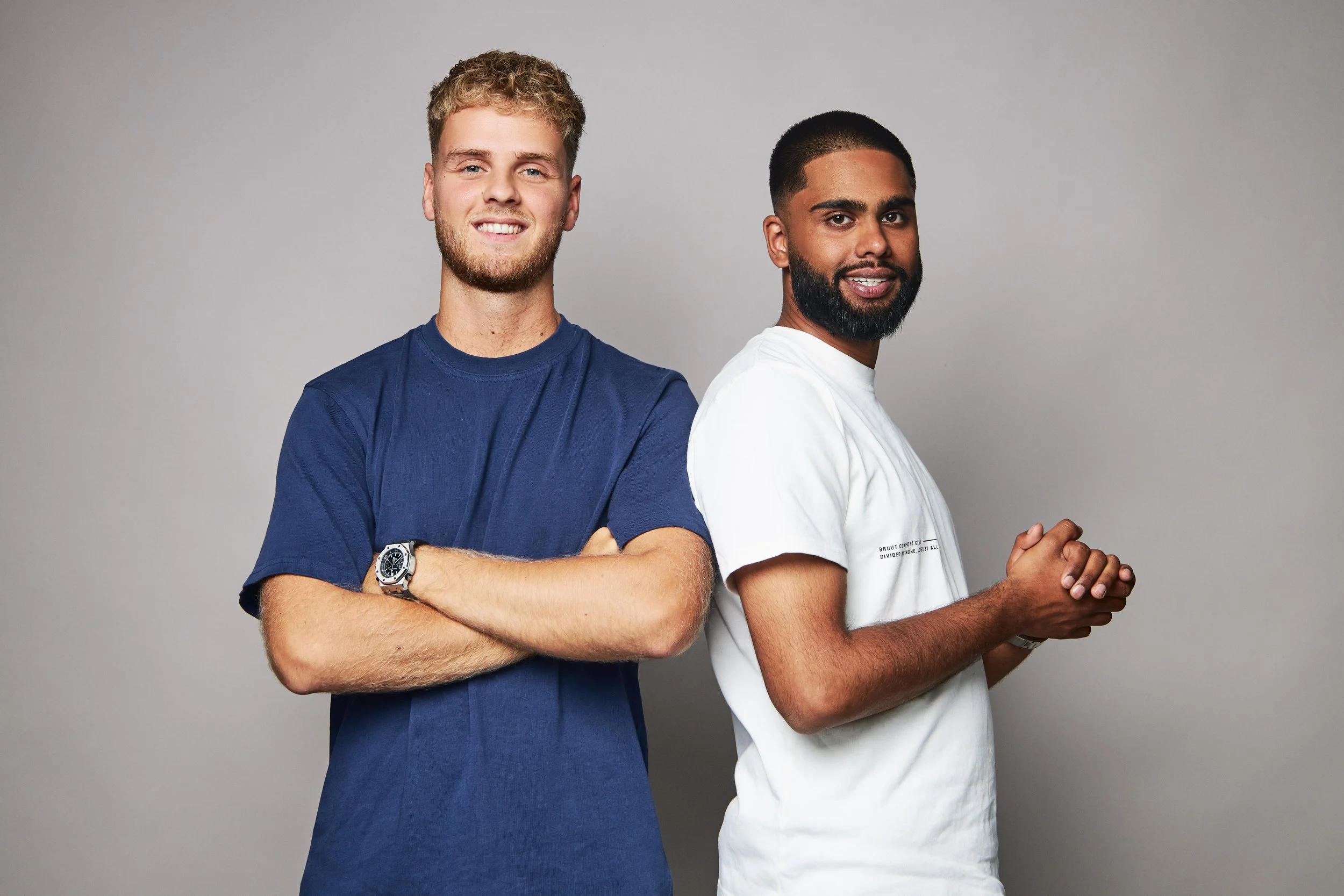 Two young men standing back-to-back against a plain gray background, smiling and clasping their hands in front of them.